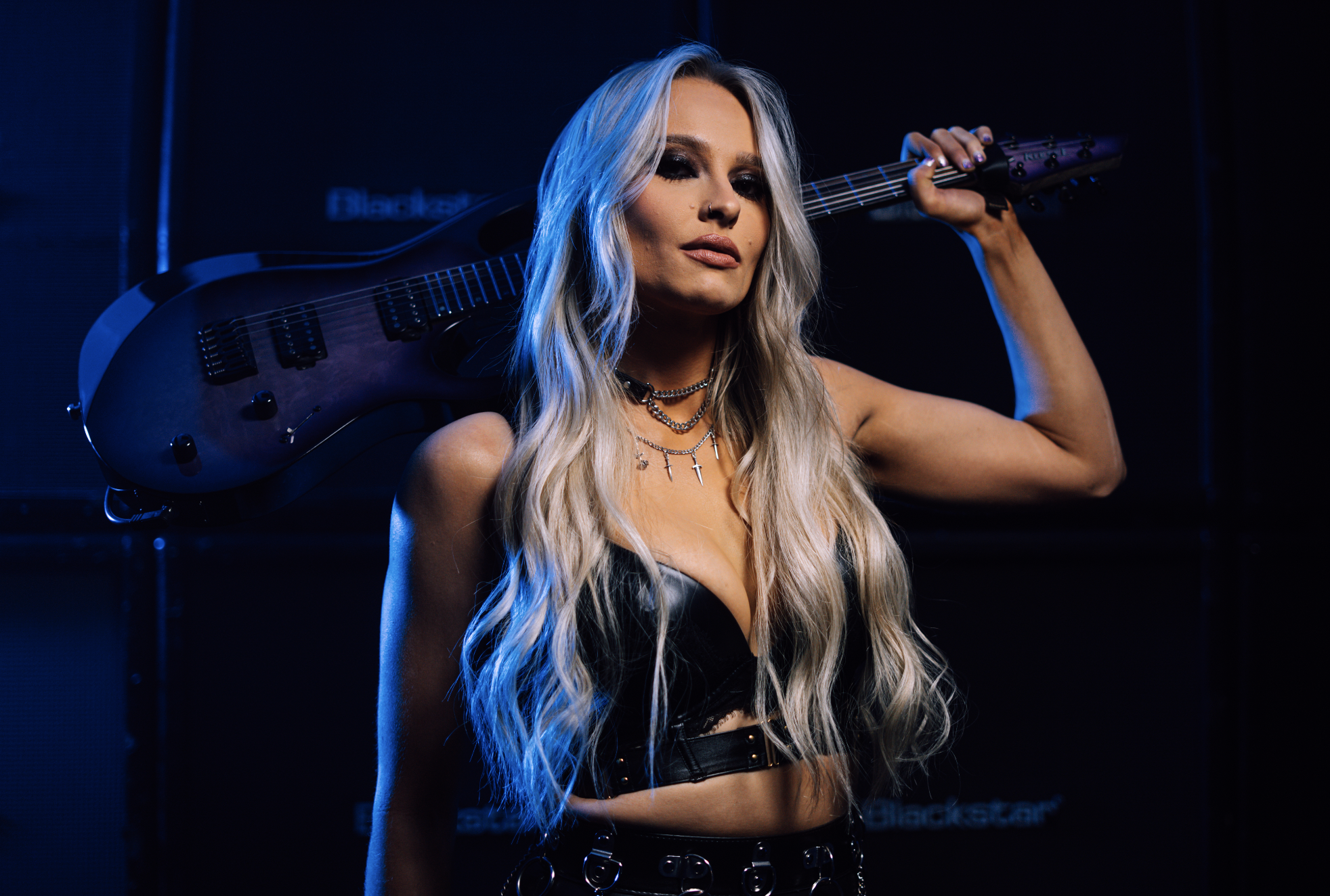 A woman with long, wavy blonde hair, wearing dark eye makeup and a black leather top, holds an electric guitar over her shoulder in a dark setting with blue lighting.