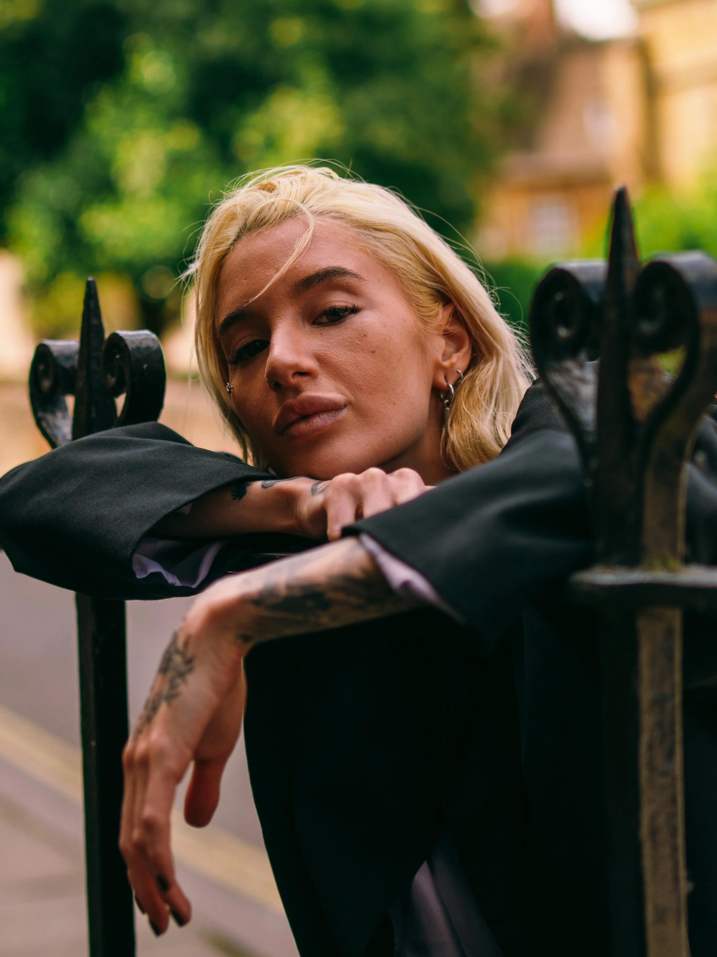A woman with platinum blonde hair and earrings leaning on a black metal fence outdoors with greenery and a building in the background.