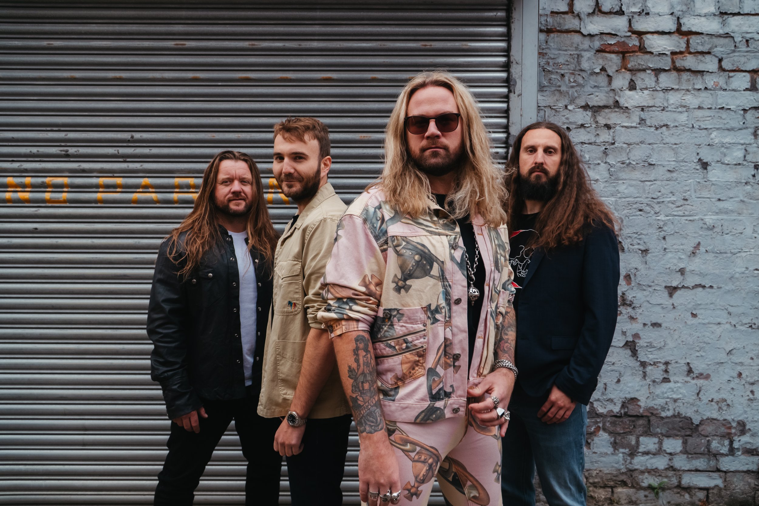 A band of four men standing in front of a corrugated metal garage door and a brick wall. The man in the center has long blond hair, sunglasses, and is wearing a colorful patterned jacket and shorts. The others have long hair and casual clothing.