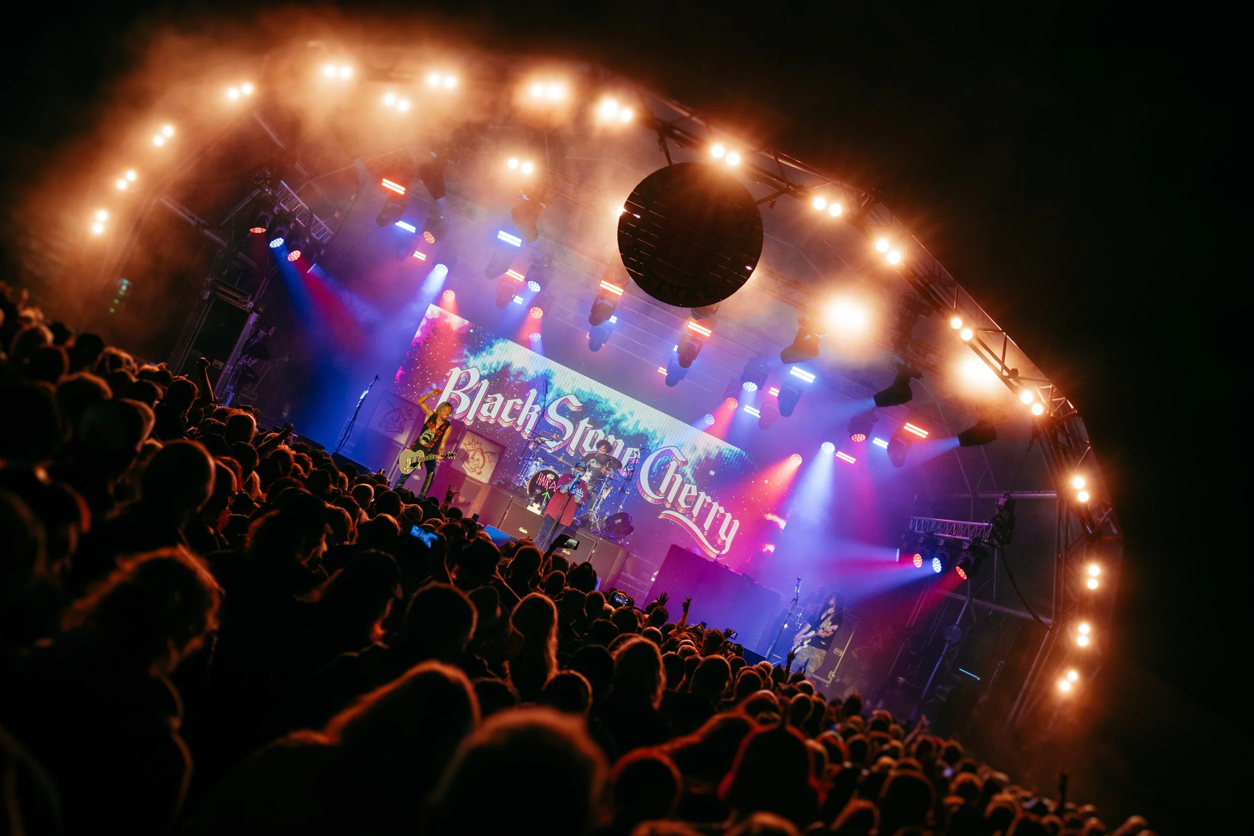 Concert stage with band performing, large crowd in front, colorful lights, and electronic display reading 'Black Star Cherry'.