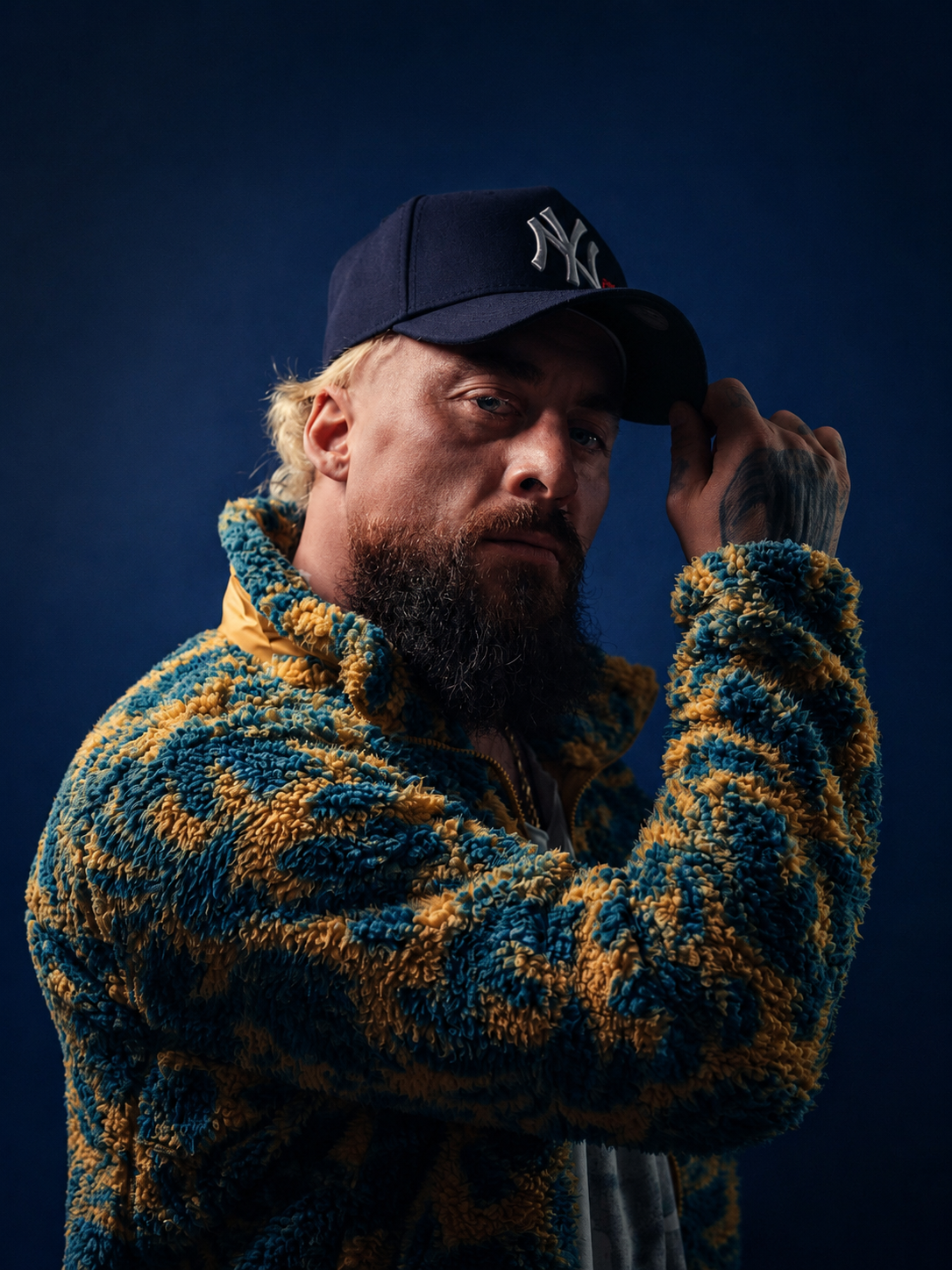 A man with blonde hair, a beard, and tattoos wears a dark blue baseball cap with the New York Yankees logo and a multicolored, textured fleece jacket, posing against a dark blue background.