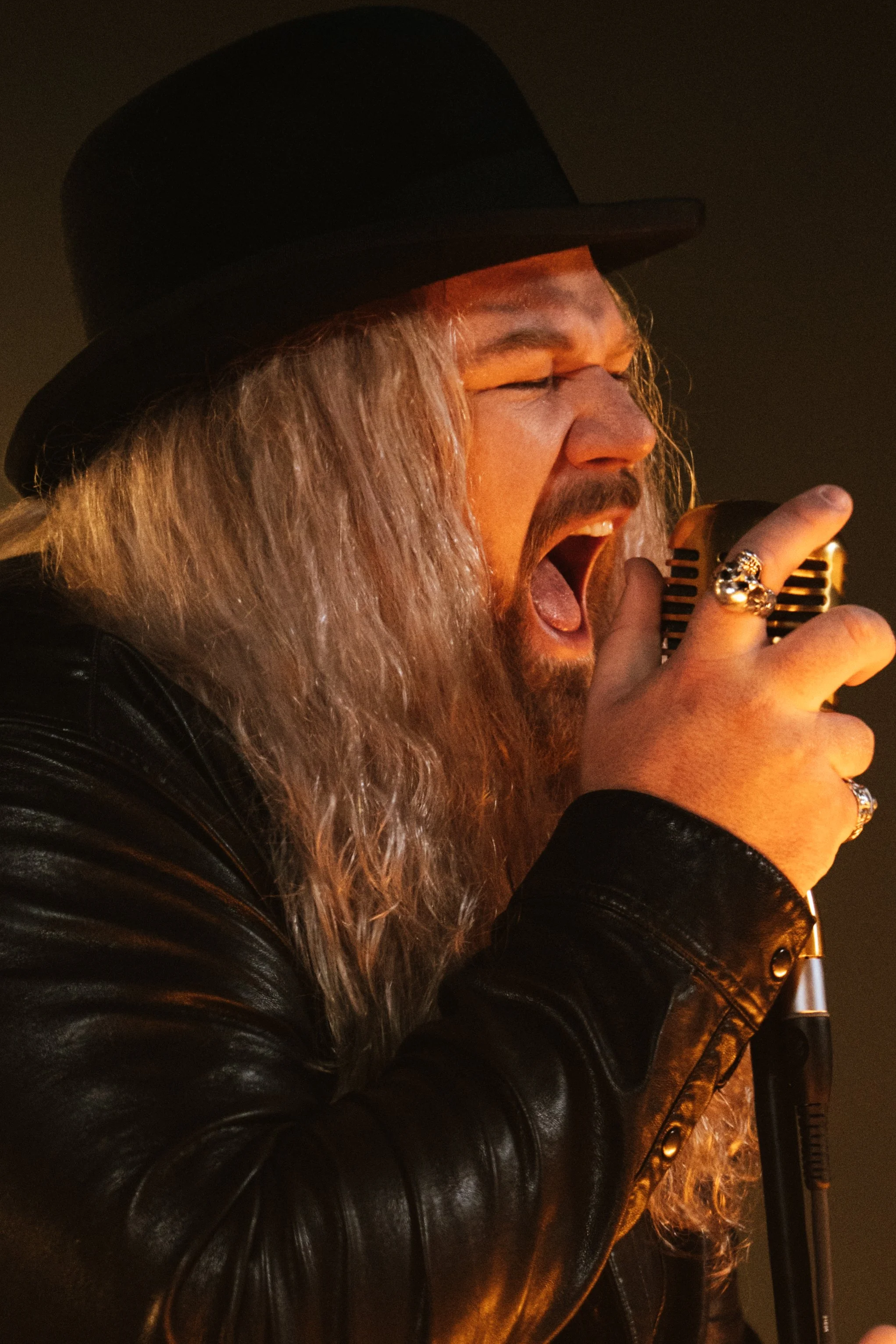 A man with long gray hair, beard, and mustache wearing a black hat and leather jacket, singing passionately into a vintage microphone.