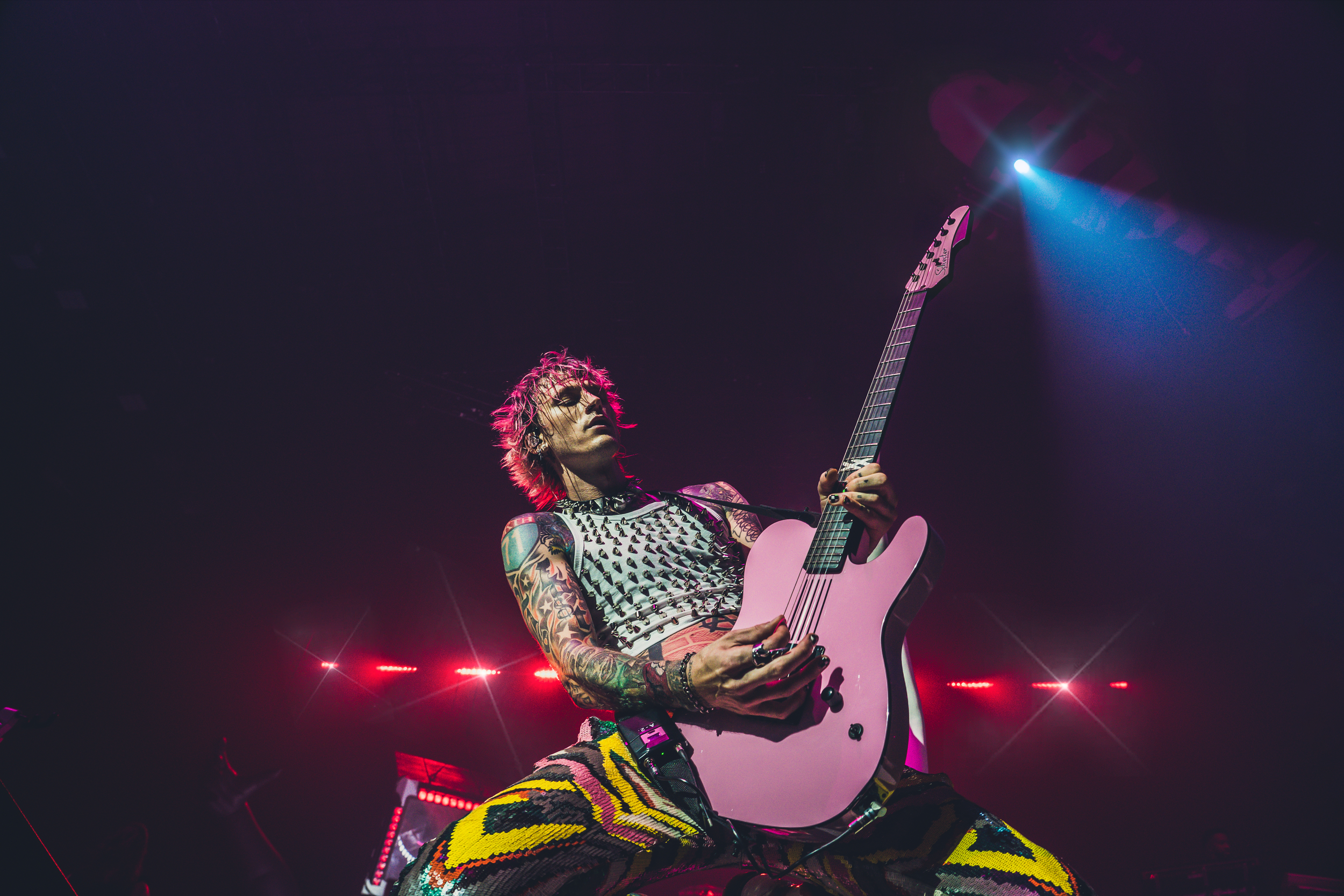 A guitarist with tattoos wearing a studded top and colorful patterned pants performs on stage, illuminated by vibrant purple and red stage lights.