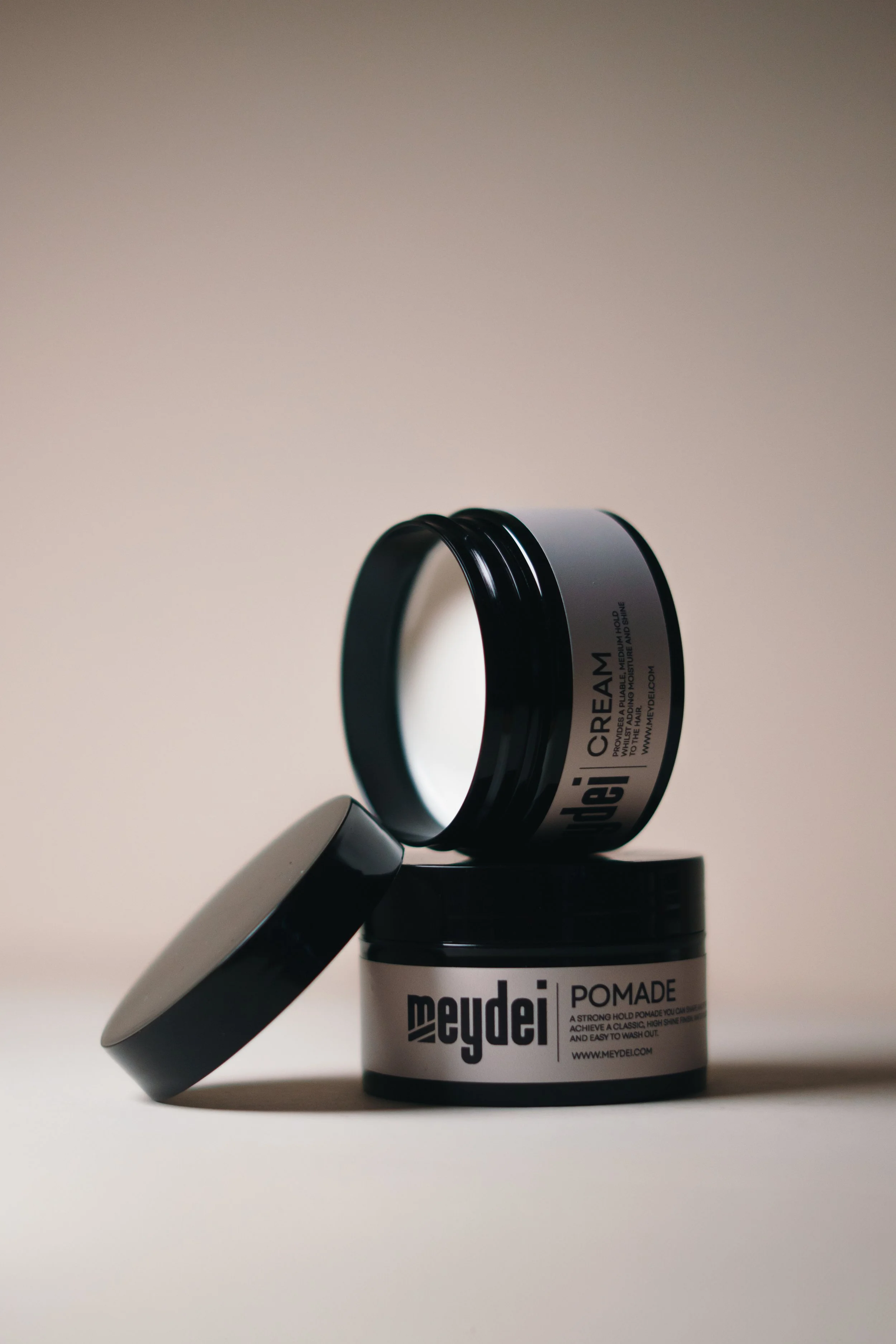 Two black and white jars of hair pomade, one open and one closed, placed on a light surface against a plain light background.