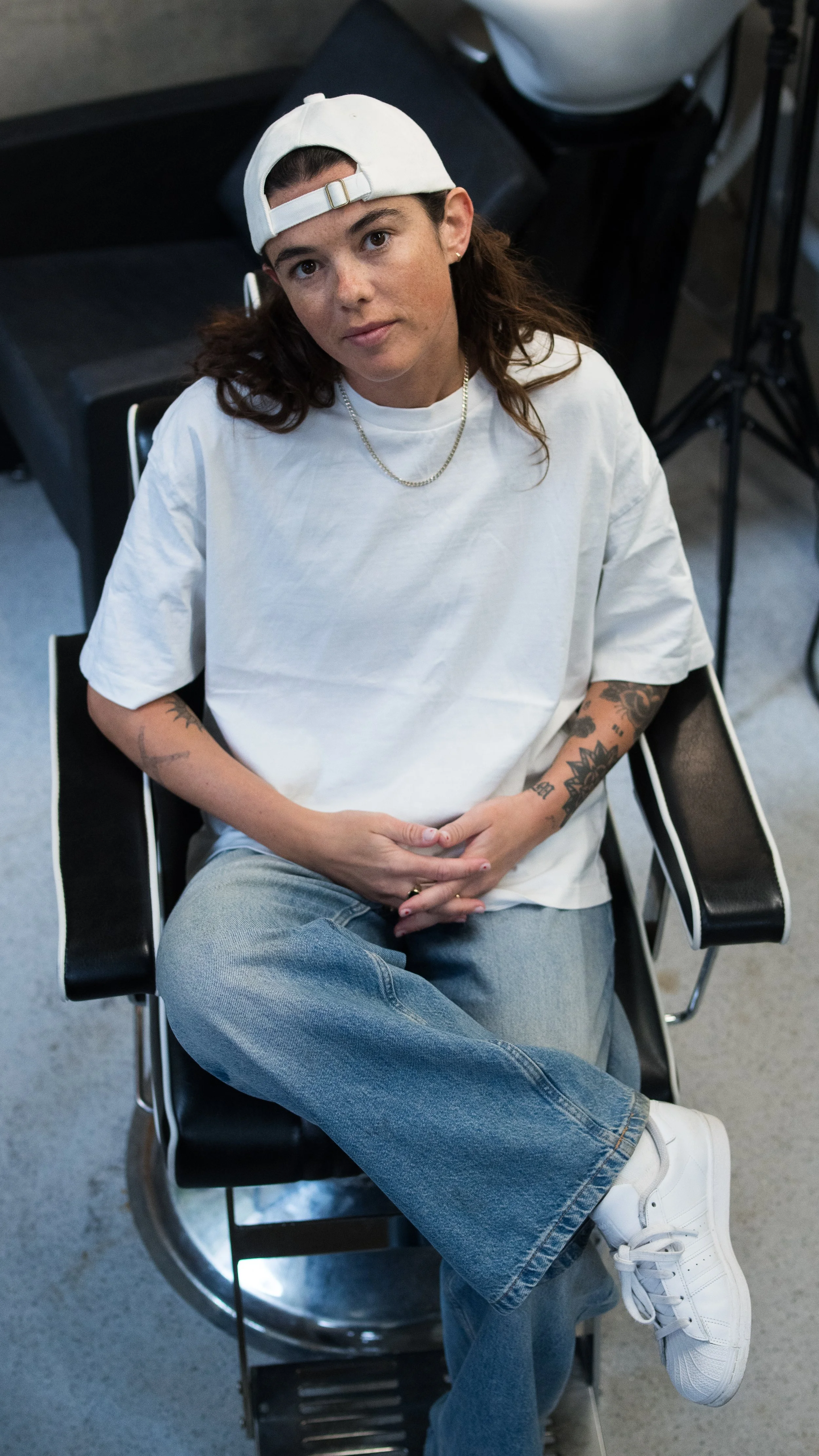 A young woman with tattoos on her arms, wearing a white t-shirt, jeans, and white sneakers, sitting in a black salon chair with her legs crossed and looking up at the camera.