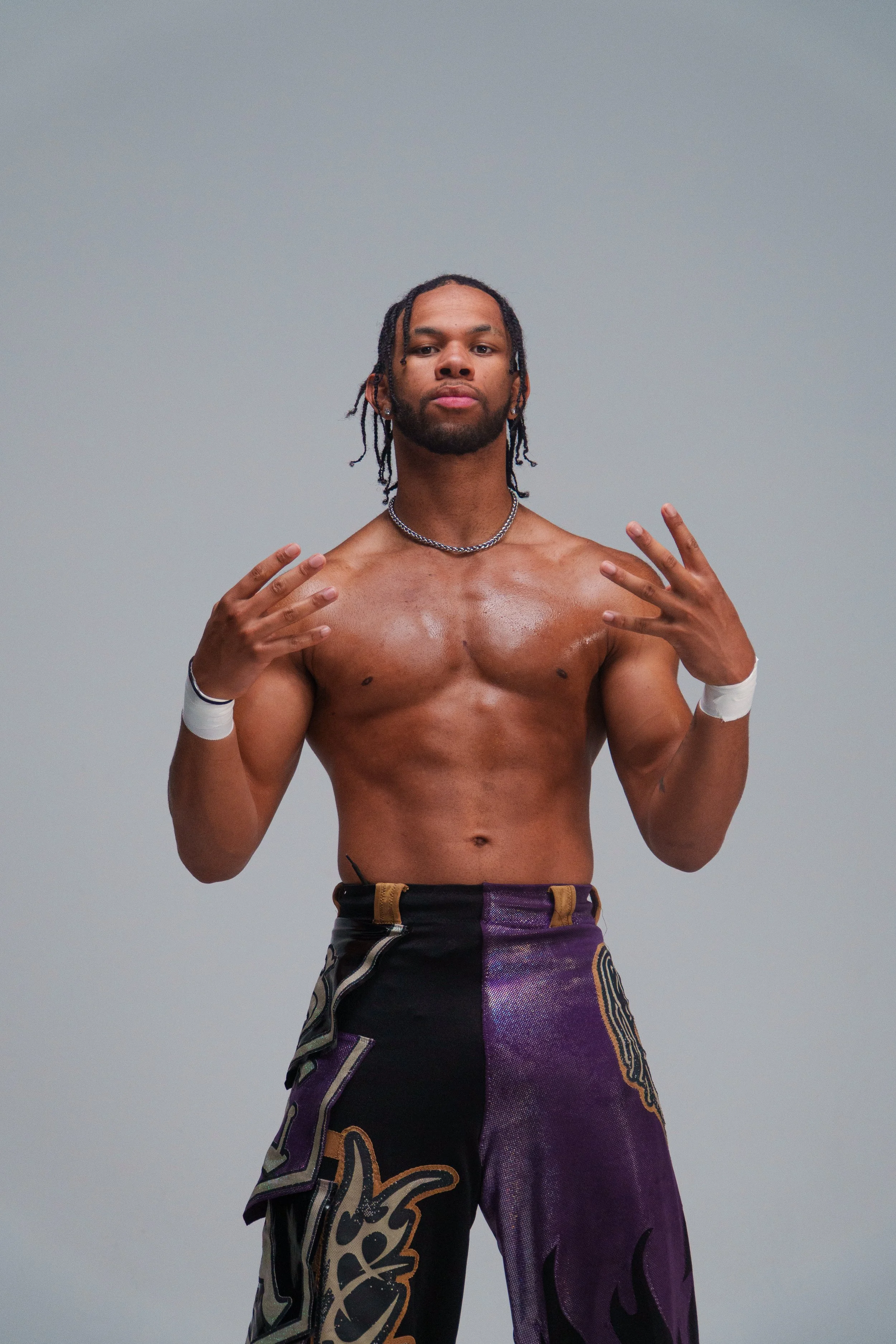 A shirtless man with dreadlocked hair, wearing a chain necklace, wristbands, and colorful pants, making a gesture with both hands against a plain gray background.