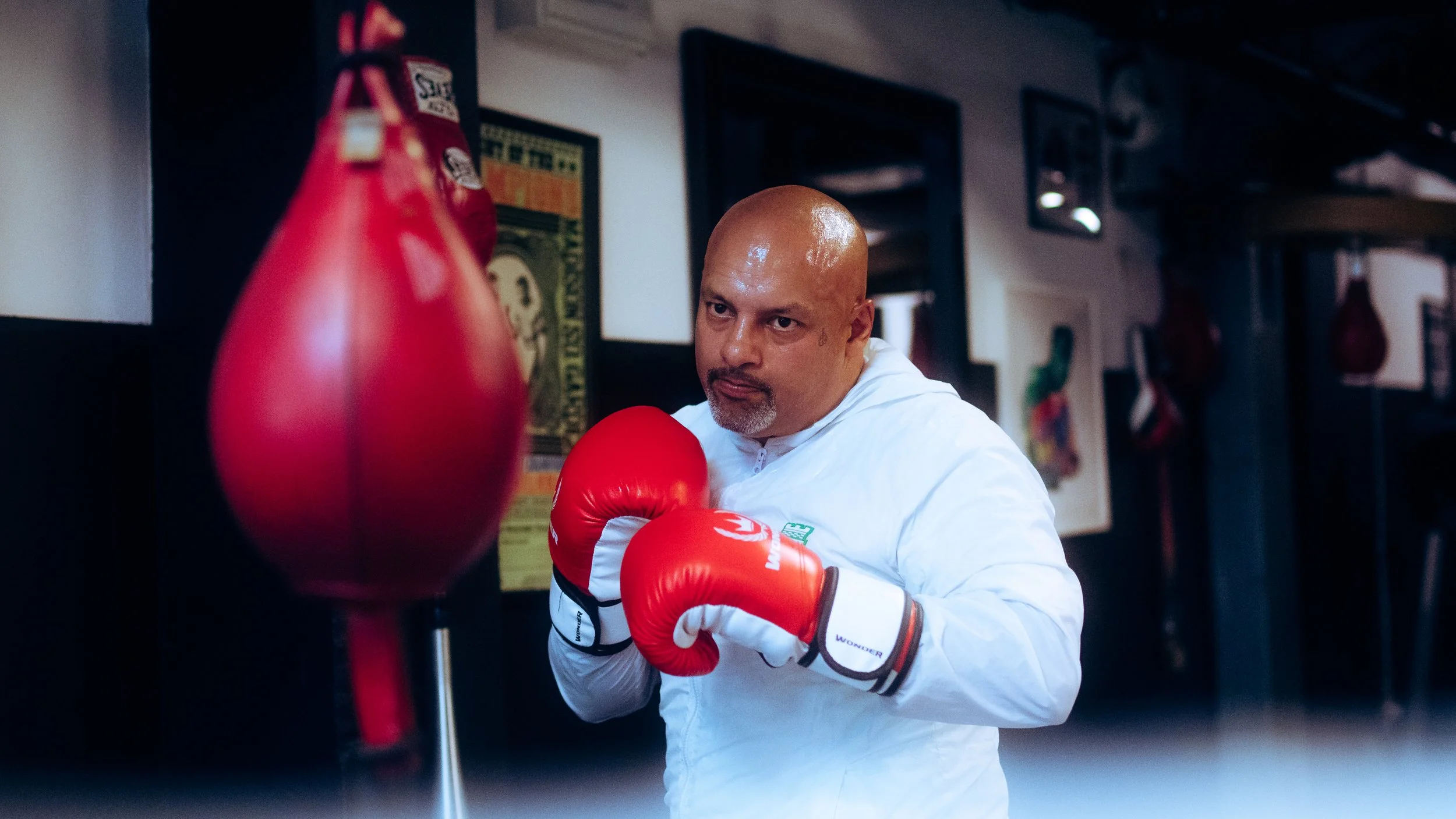 A man with a shaved head and goatee in a white shirt throws a punch at a red punching bag in a boxing gym.