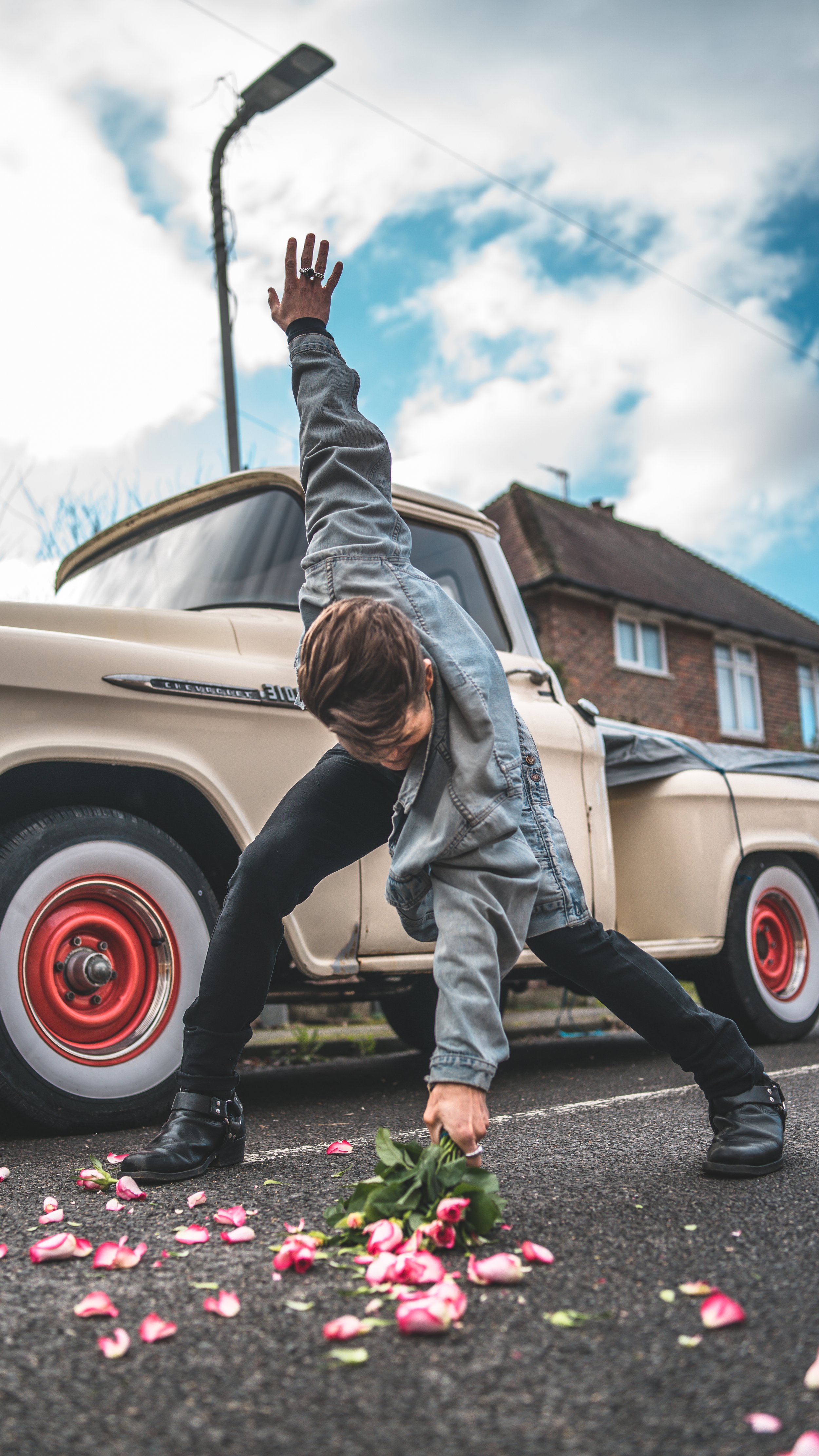A woman in black boots and a denim jacket bends down to pick up pink and white roses from the road, with a vintage beige pickup truck and a house with a brick wall and a chimney in the background, under a cloudy sky.