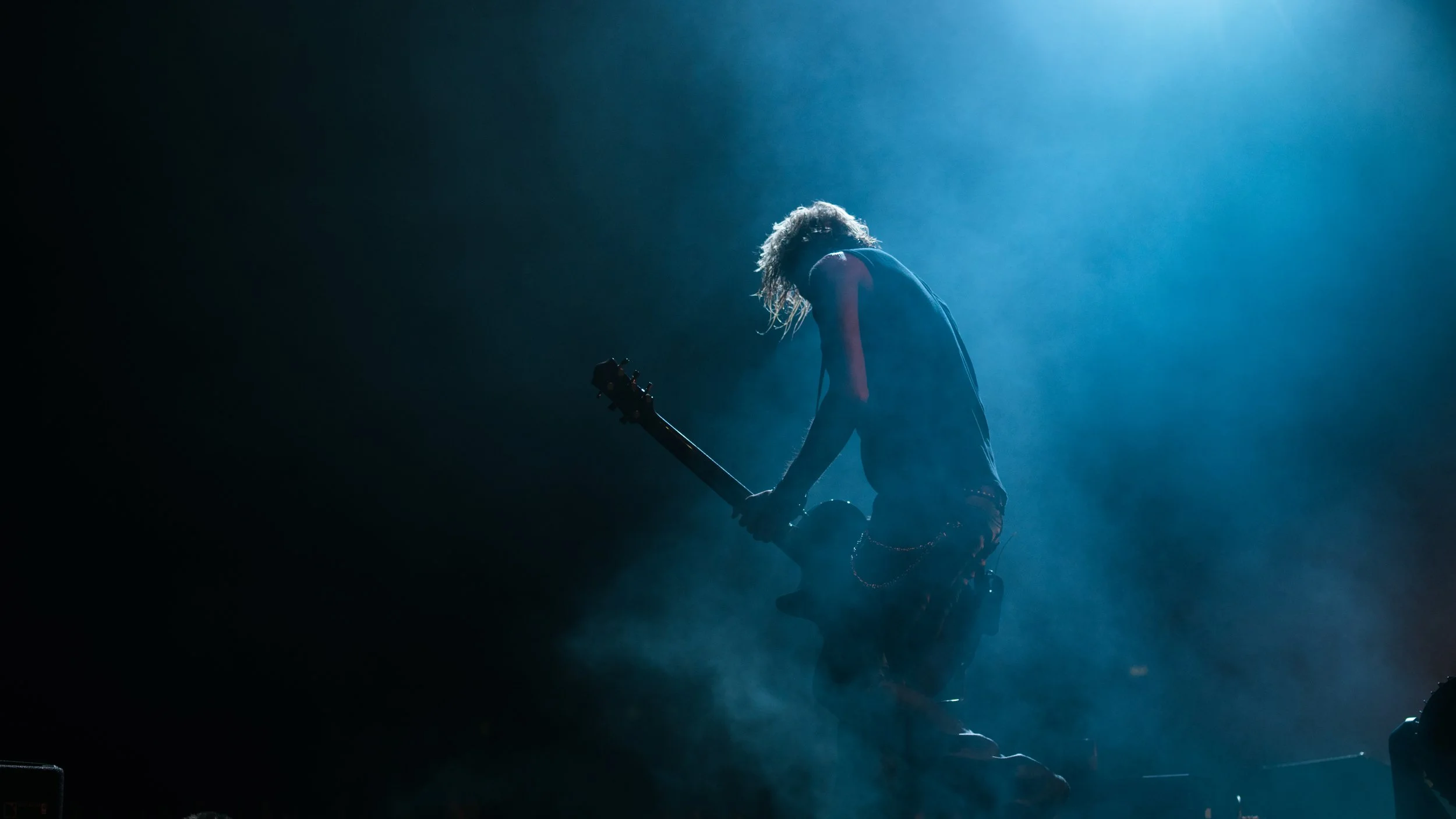 Silhouette of a musician kneeling with a guitar on a stage illuminated by a bright blue spotlight, with smoke or fog around.