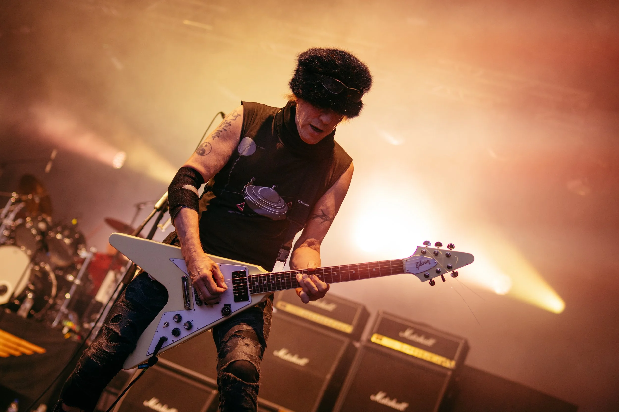 A guitarist wearing a fluffy black hat, sunglasses, and a sleeveless shirt playing a white Gibson electric guitar on stage with amplifiers in the background.