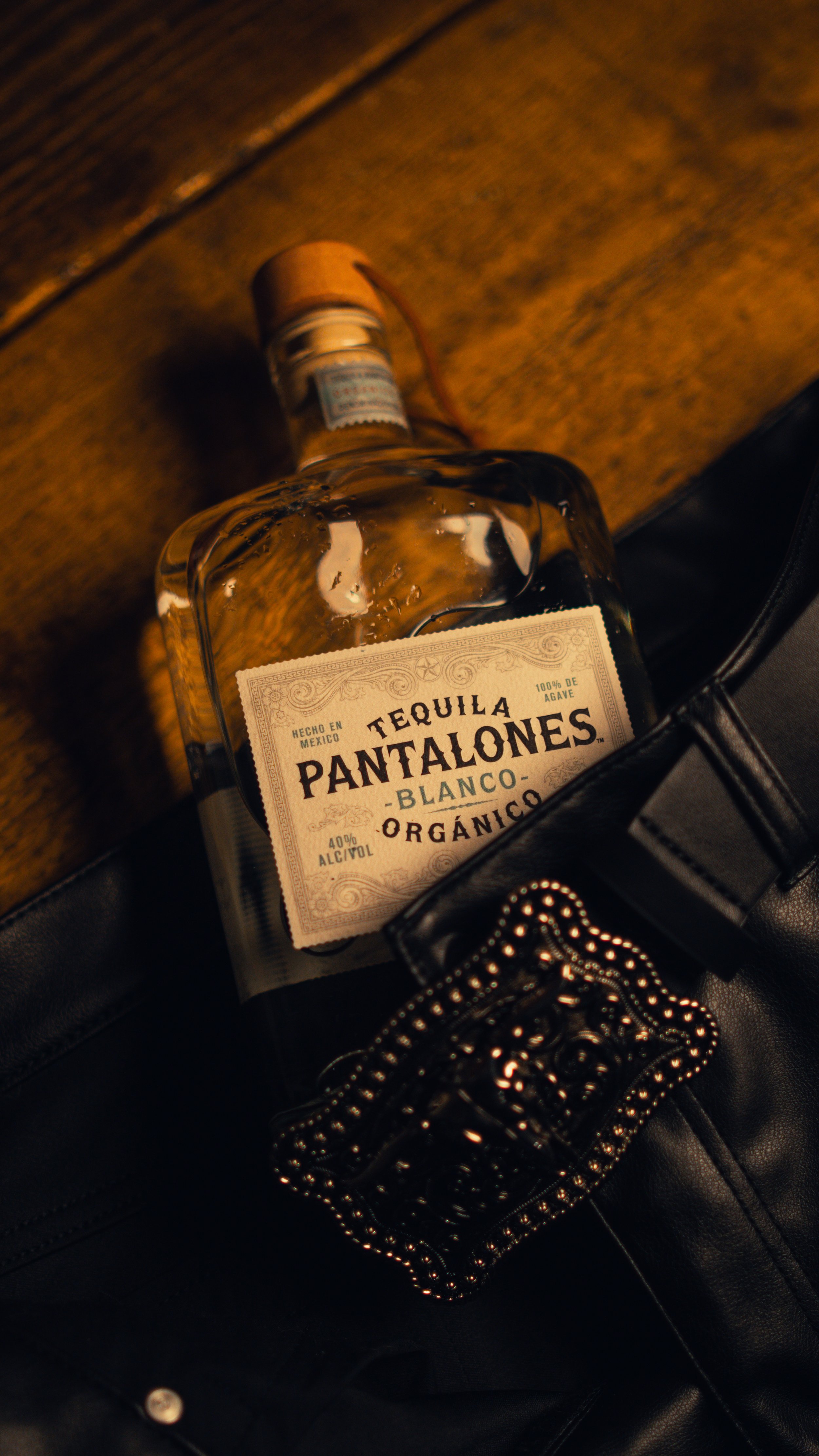 A clear glass bottle of Tequila Pantalones Blanco Organico resting on a black leather bag with a decorative chain, placed on a wooden surface.