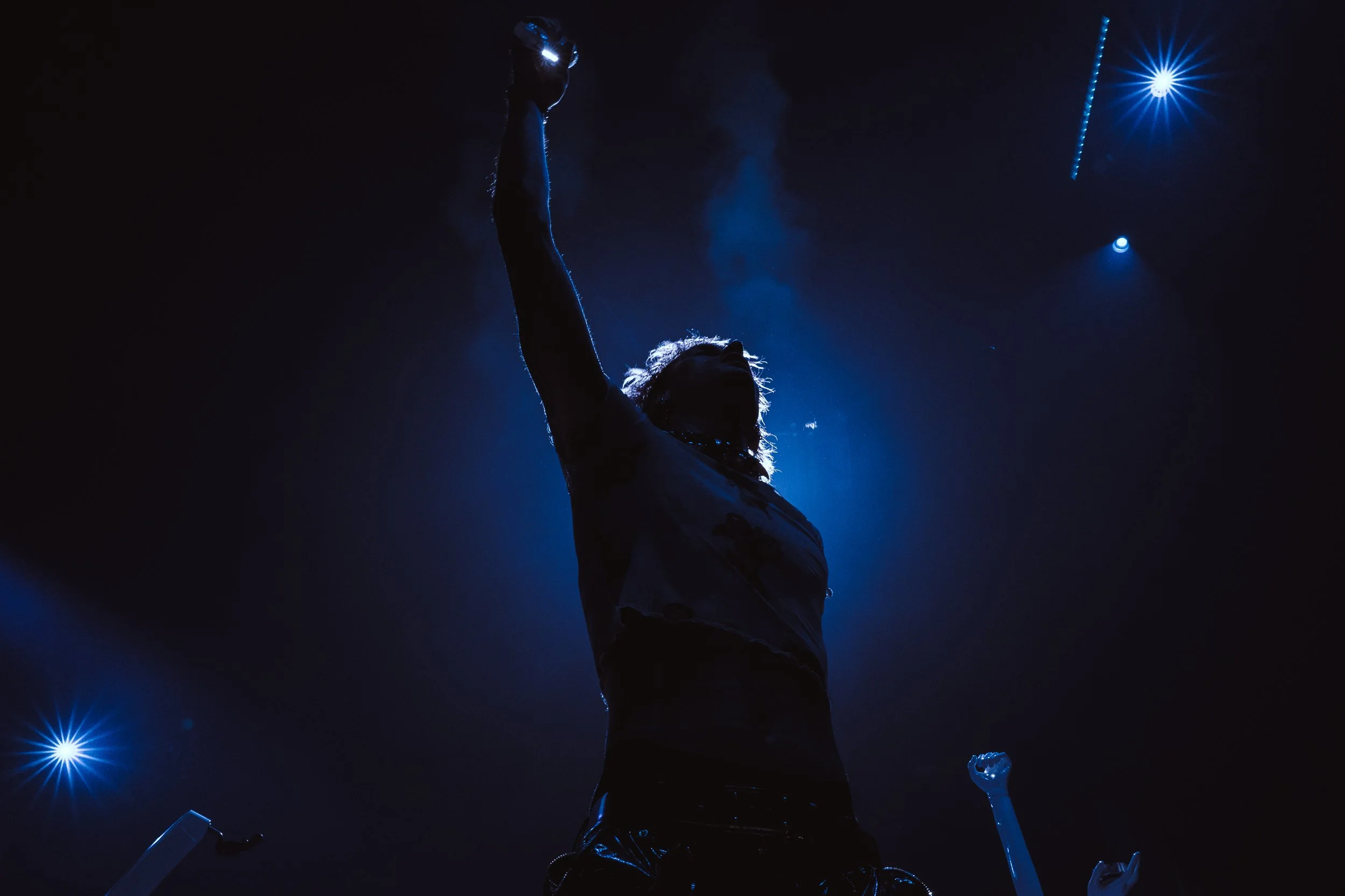 Silhouette of a person lifting their fist in the air on a dark stage with blue spotlight lighting and smoke.