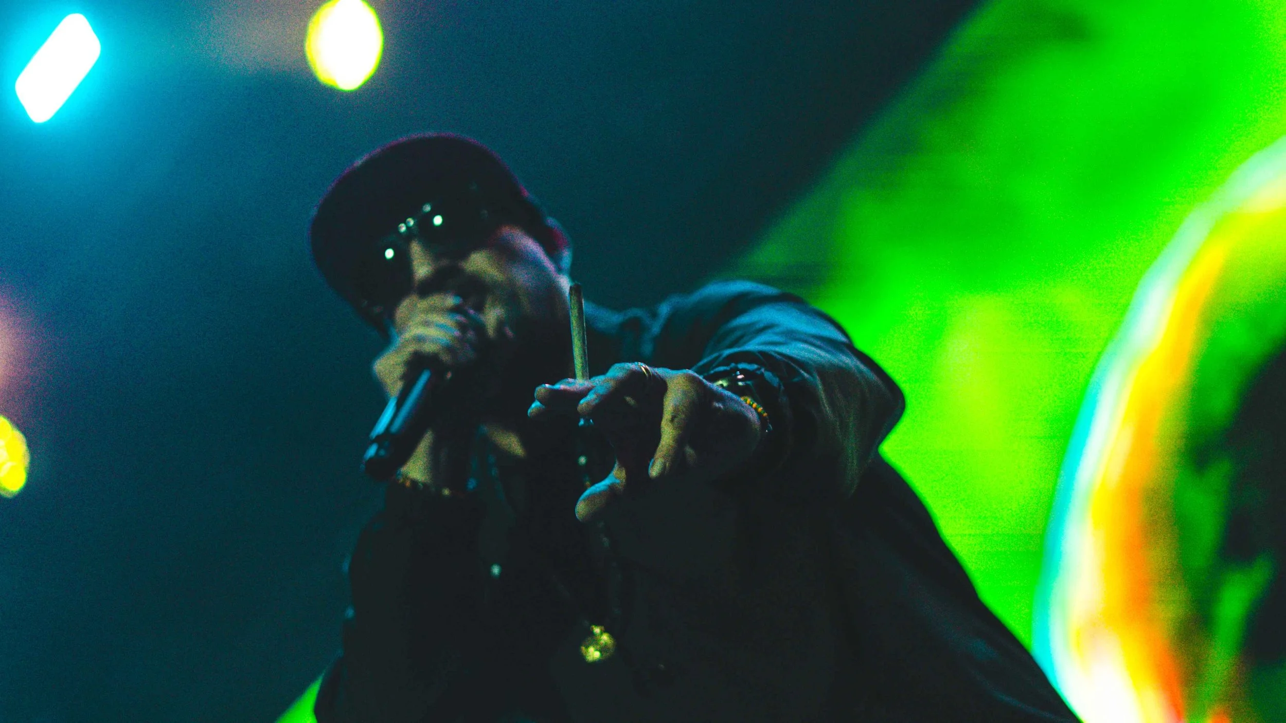 Close-up of a man wearing sunglasses and a cap, holding a microphone and pointing towards the camera on stage with colorful lights in the background.