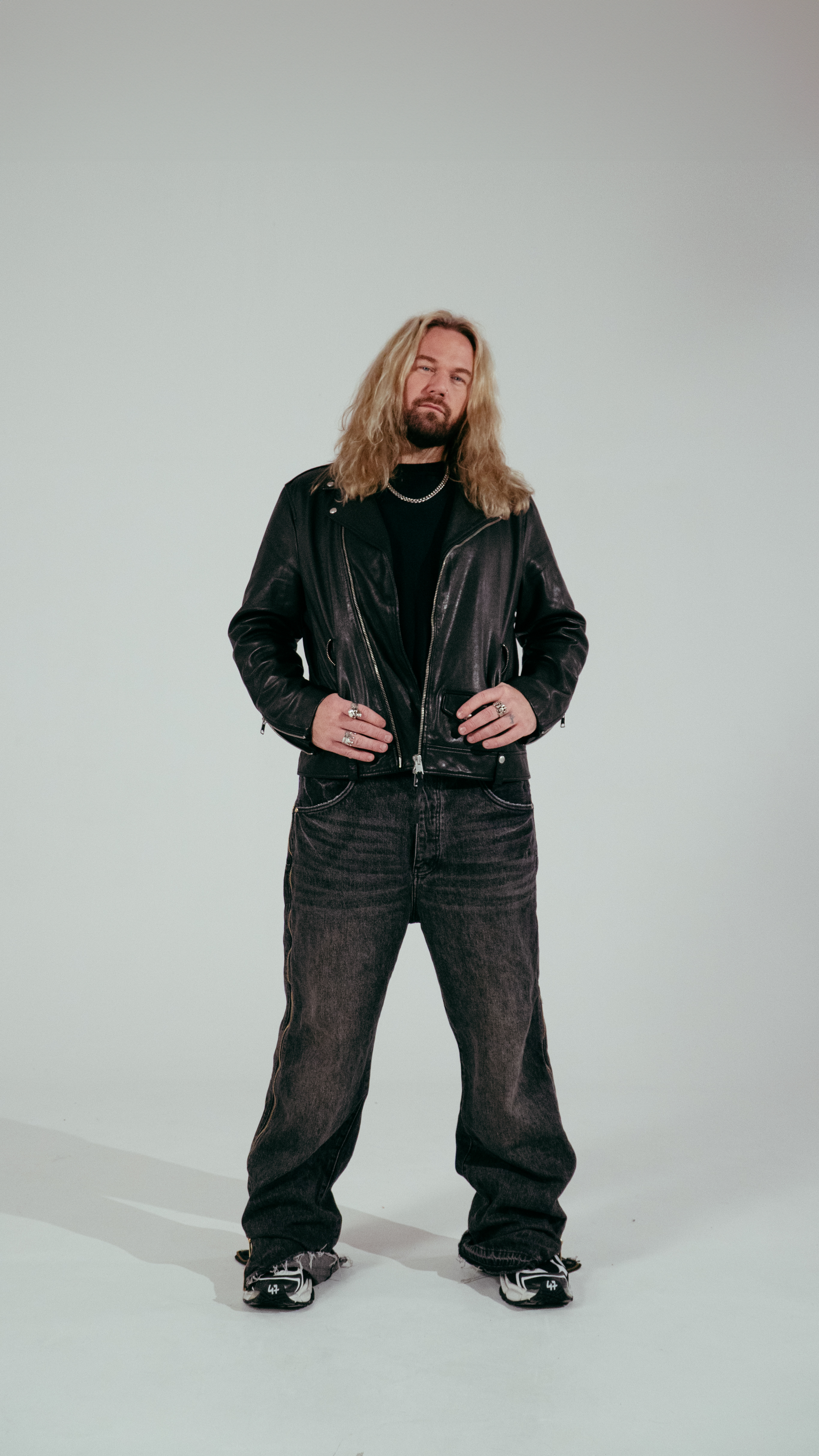 A man with long blond hair and a beard, wearing a black leather jacket, black shirt, black jeans, and sneakers, standing against a light gray background with his hands on his hips.