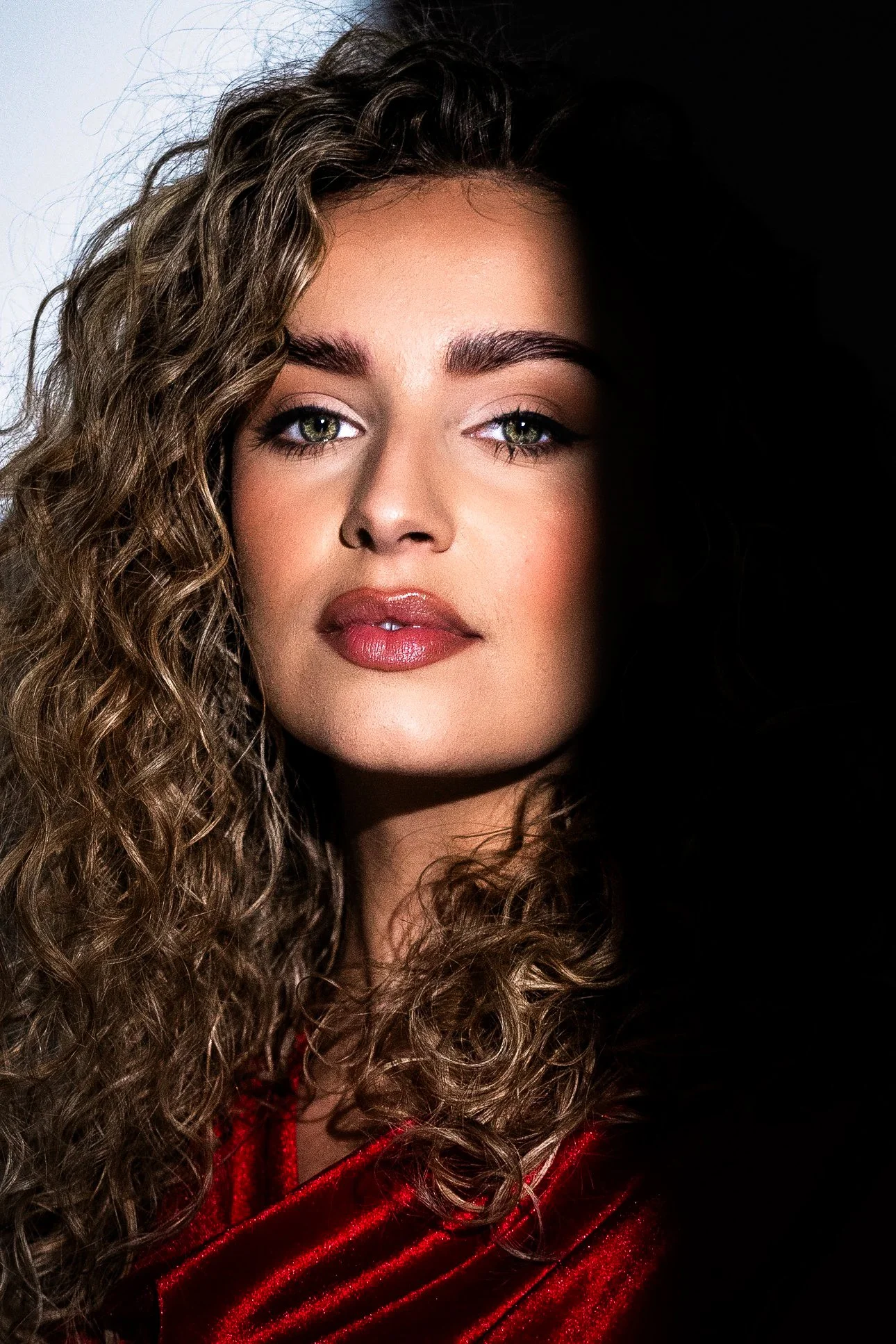 Close-up of a young woman with curly hair, light skin, and green eyes, wearing a red velvet top, with one side of her face in shadow.