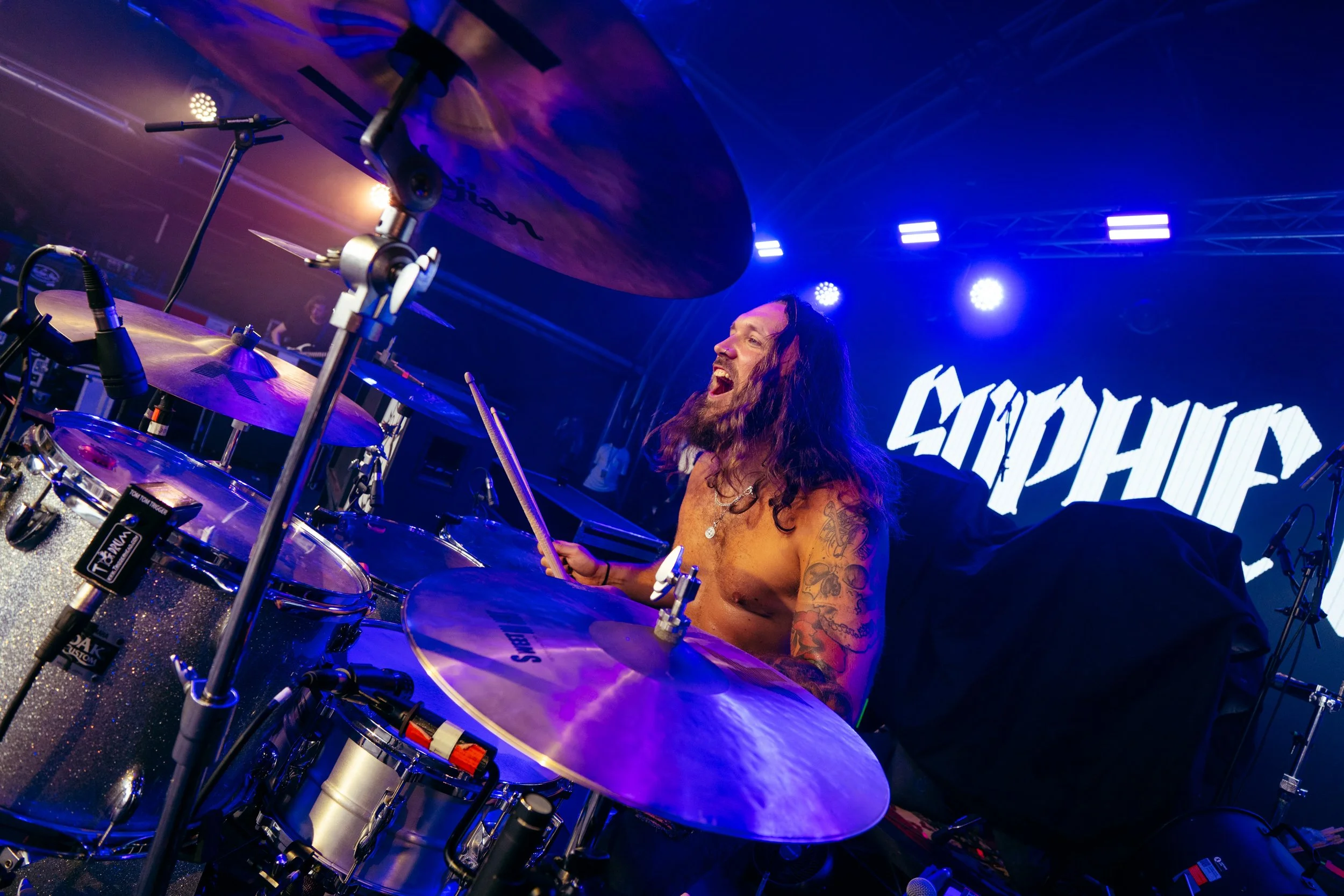 A shirtless male drummer with long hair and tattoos on his arm playing drums on stage with a big smile, under purple and blue stage lights, with a backdrop that reads 'MOPUP.'