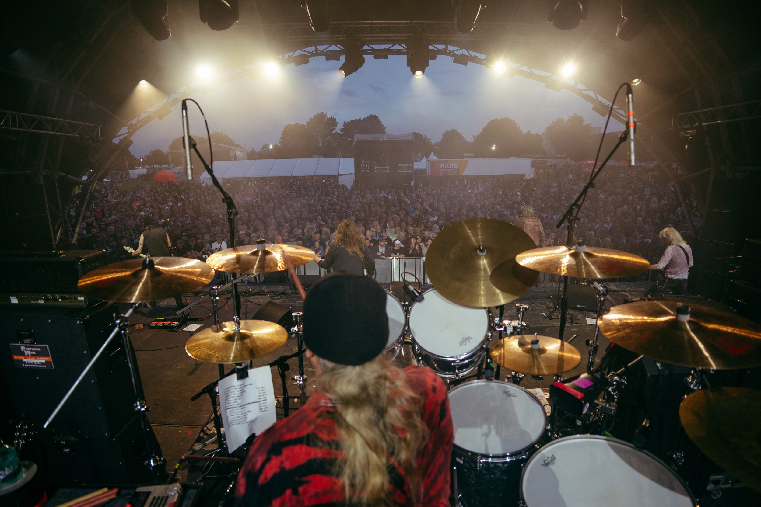 Musicians performing on stage at an outdoor concert, with a large crowd in the audience and trees in the background during dusk.