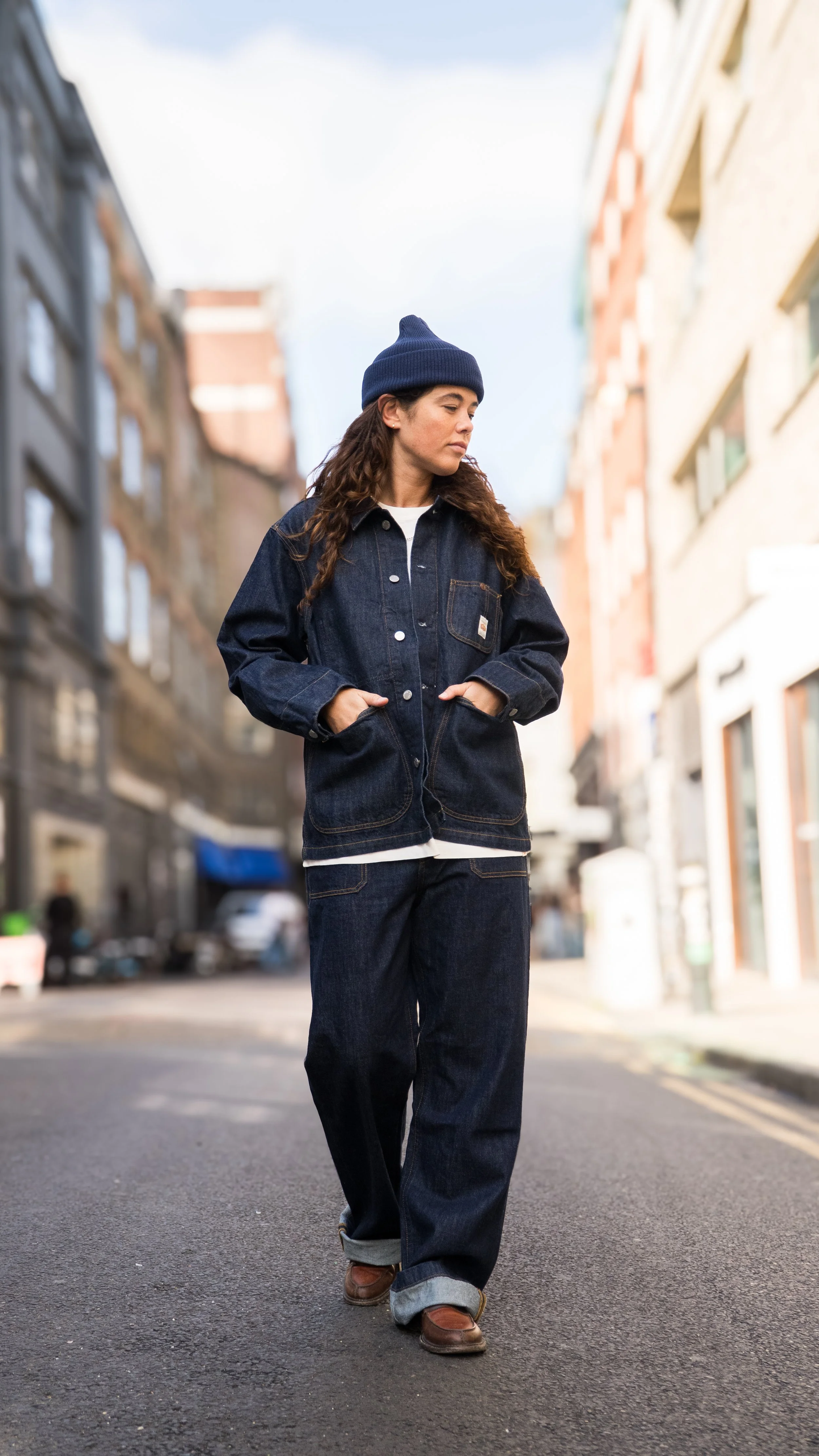 Woman walking on city street wearing denim jacket, jeans, and beanie.