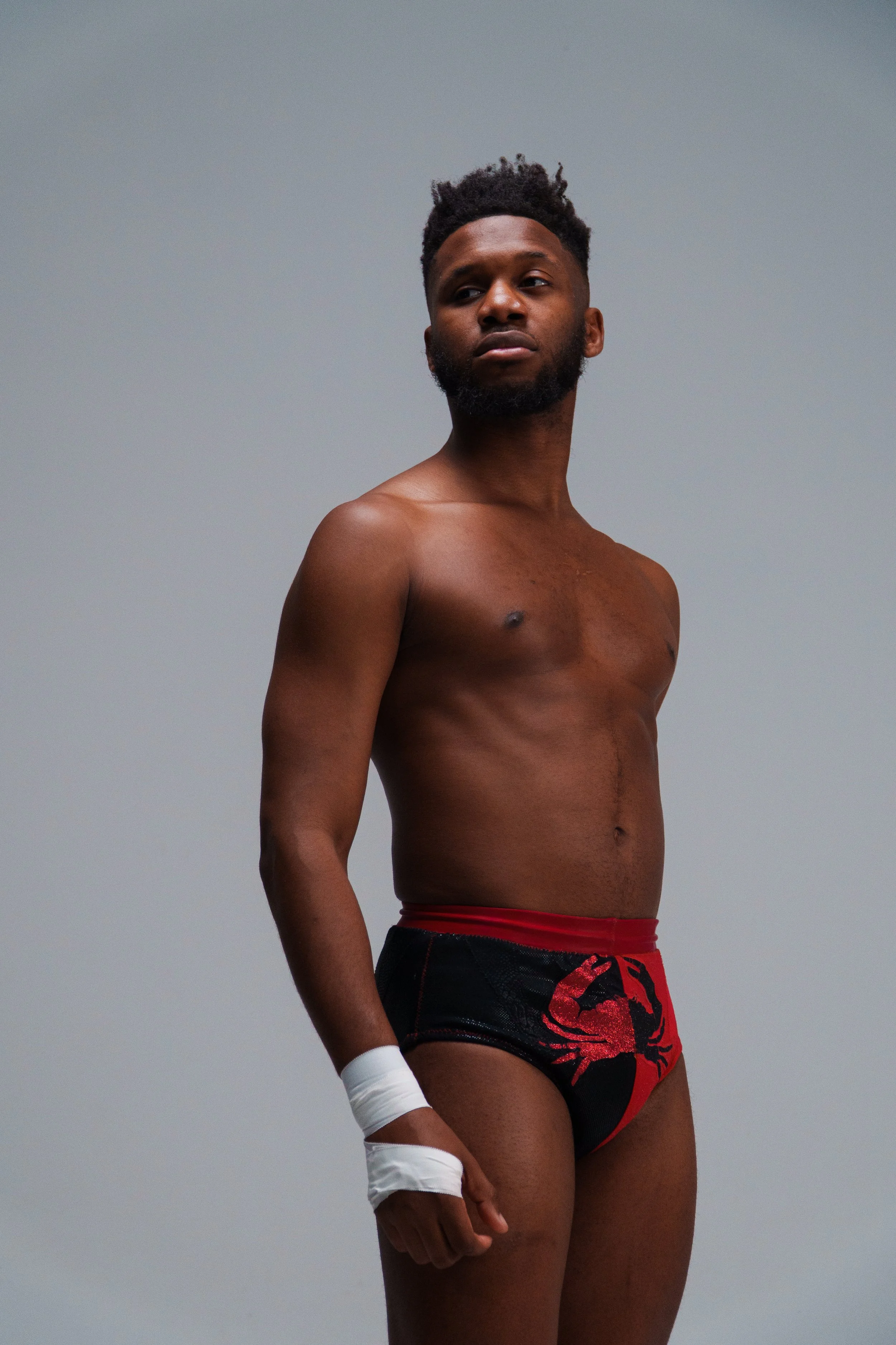 A shirtless man with a beard and short, curly hair standing against a plain gray background, wearing black and red wrestling shorts with a red scorpion design, and white wrist tape on one wrist.