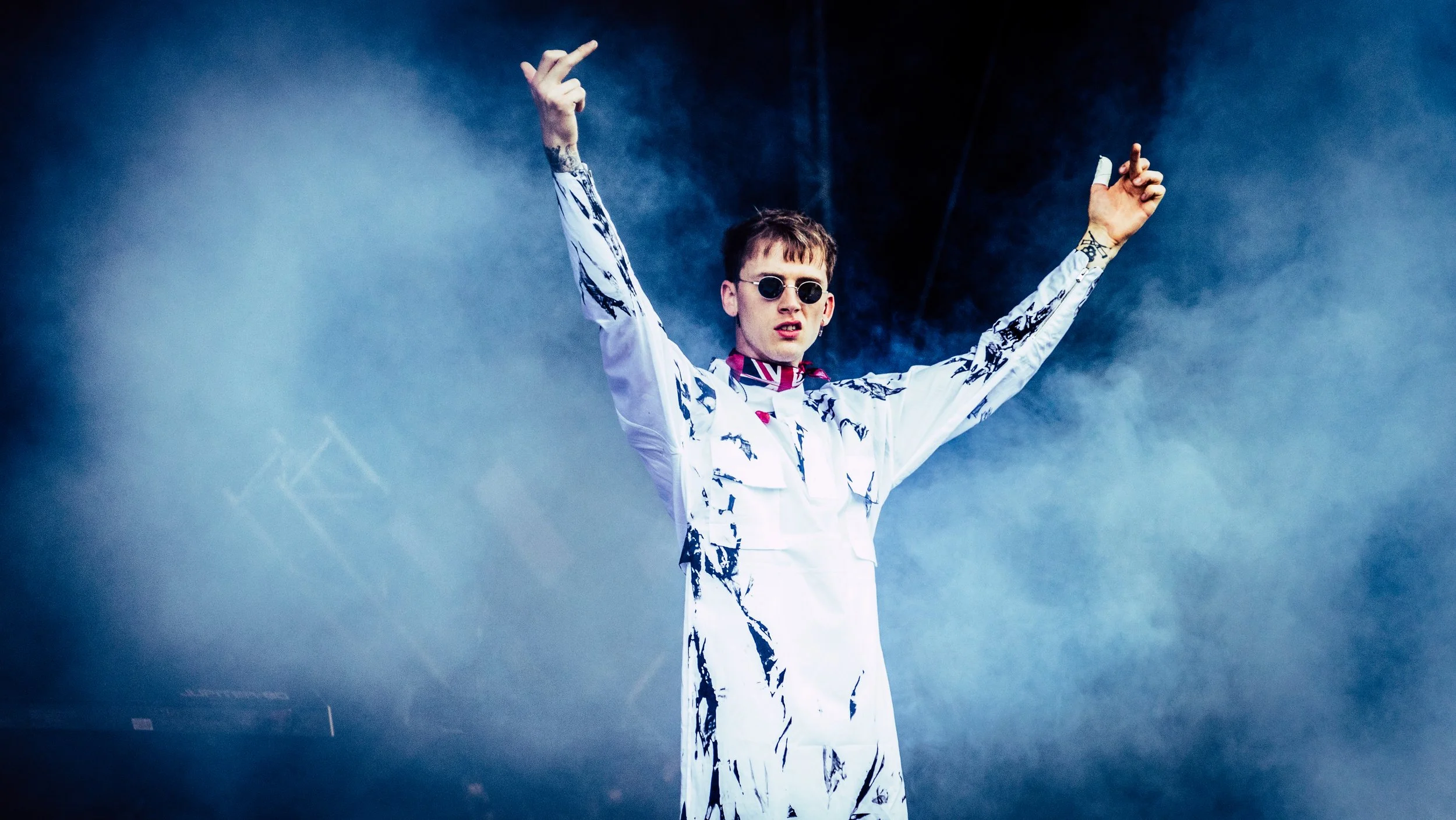 A young man with short brown hair wearing dark sunglasses and a white patterned jacket with a British flag scarf is performing on stage with arms raised.