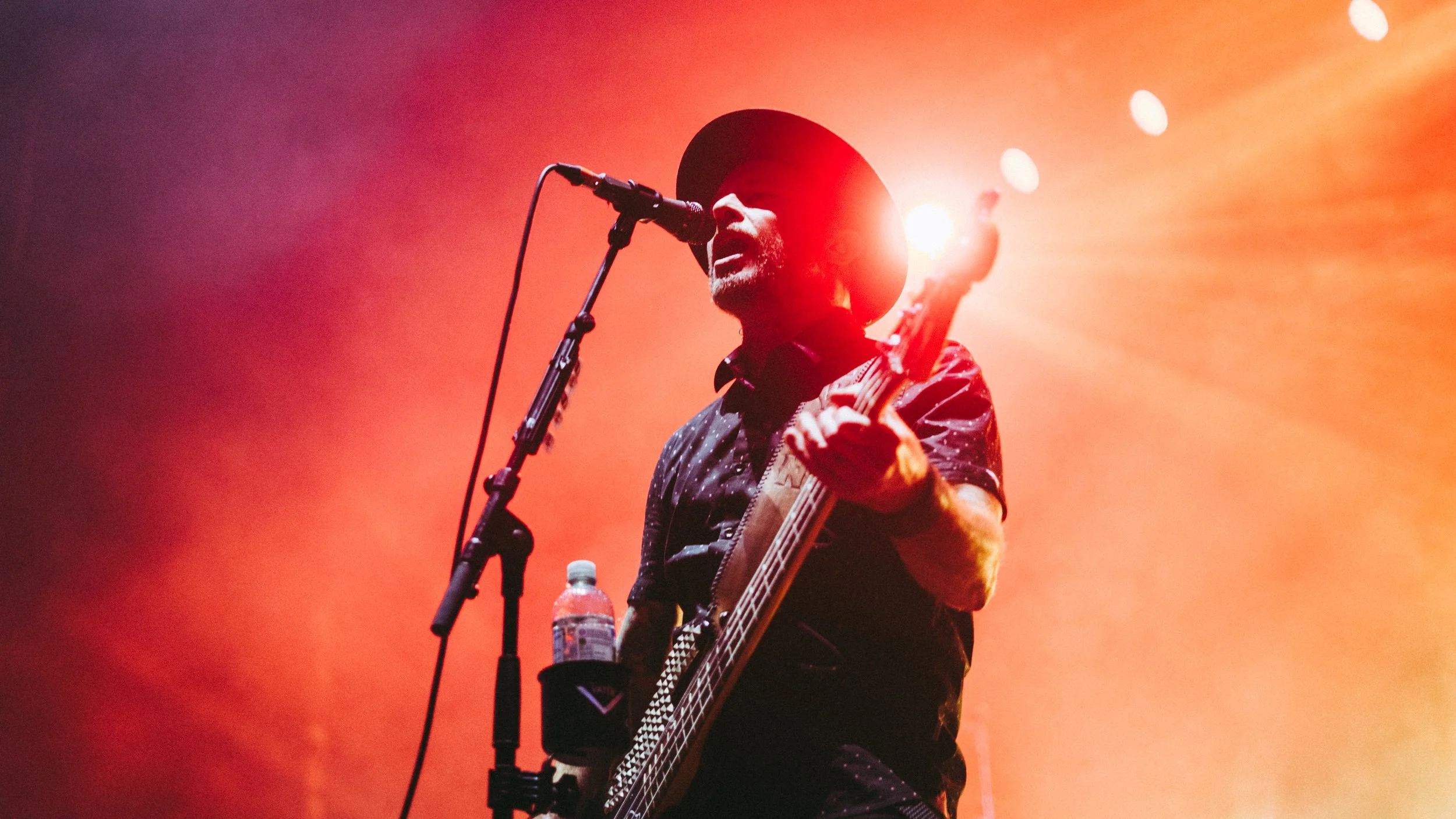 A man wearing a wide-brimmed hat and dark shirt is singing into a microphone while playing an acoustic guitar on stage, with warm orange stage lighting in the background.