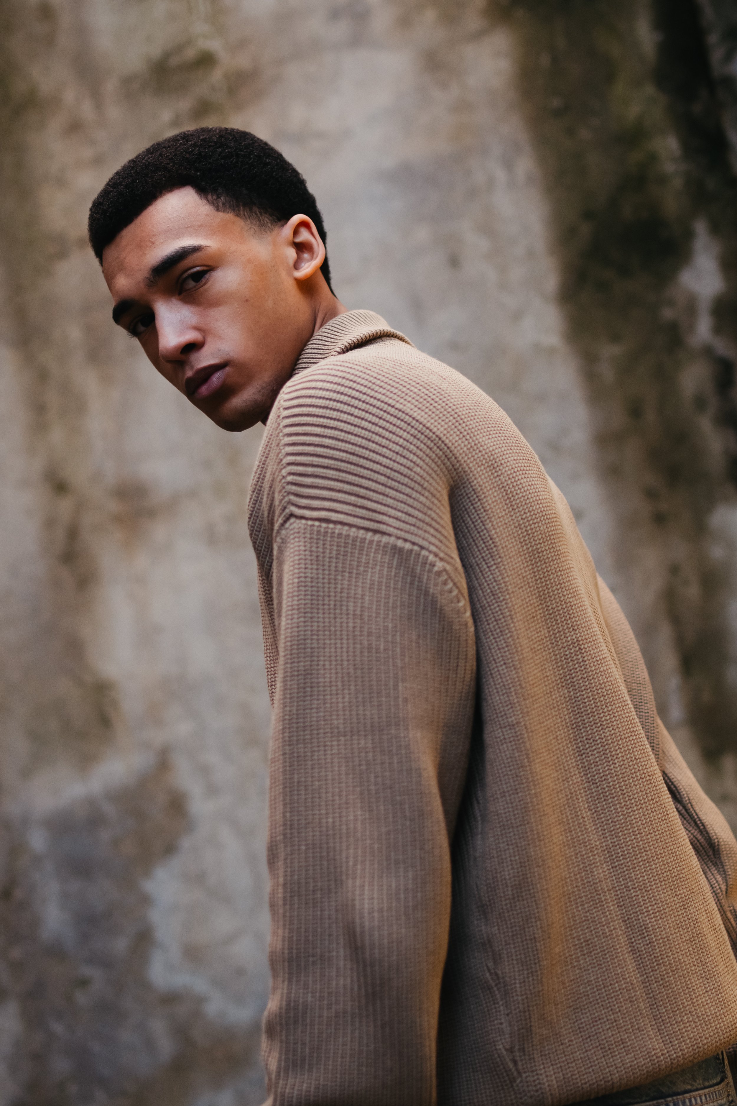 A young man with short curly black hair and dark skin stands against a textured stone wall, wearing a beige textured jacket, looking over his shoulder at the camera.