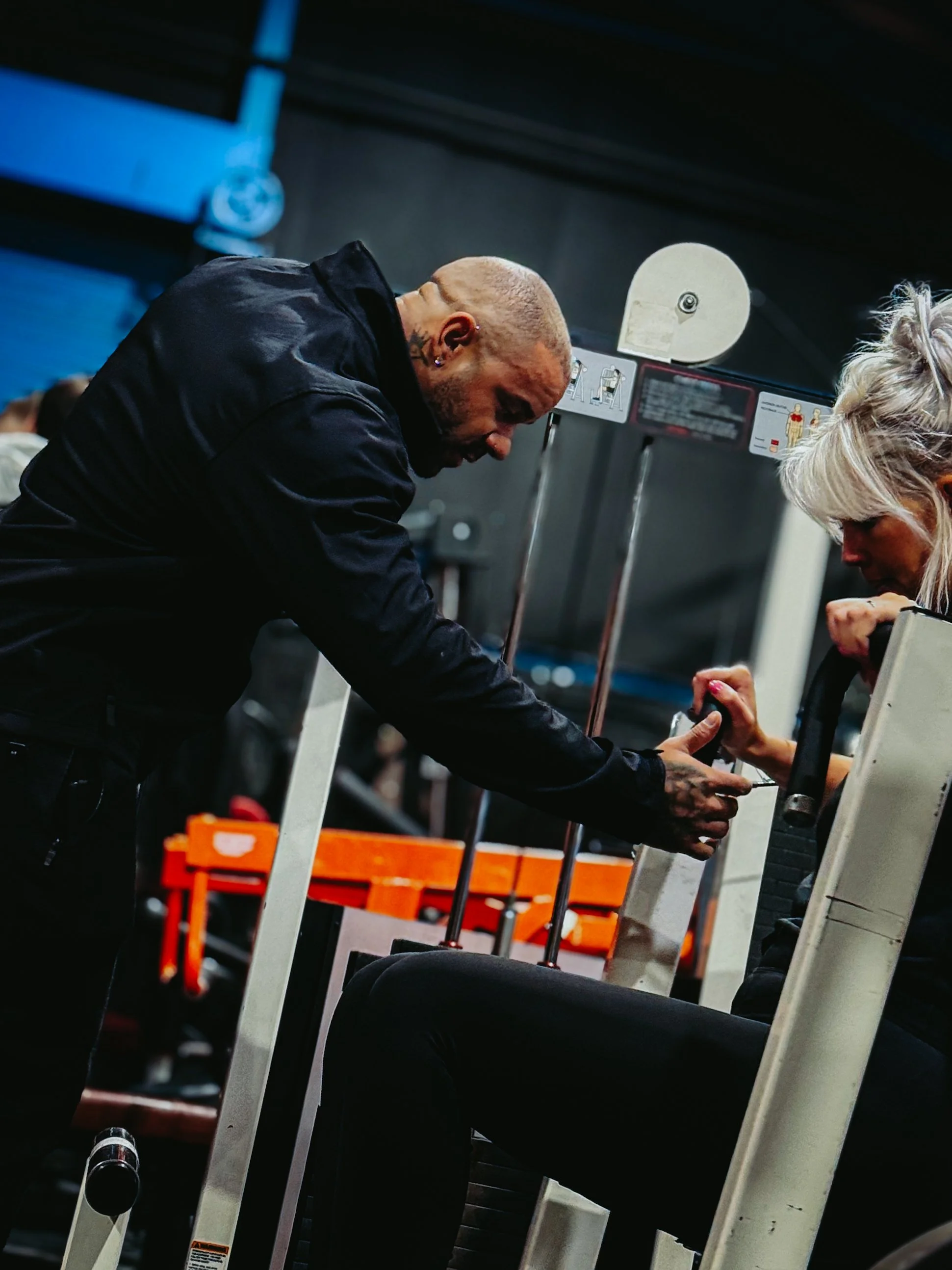 A muscular man with a mohawk hairstyle and tattoos on his arms is interacting with a woman at a gym, showing her how to use some equipment.