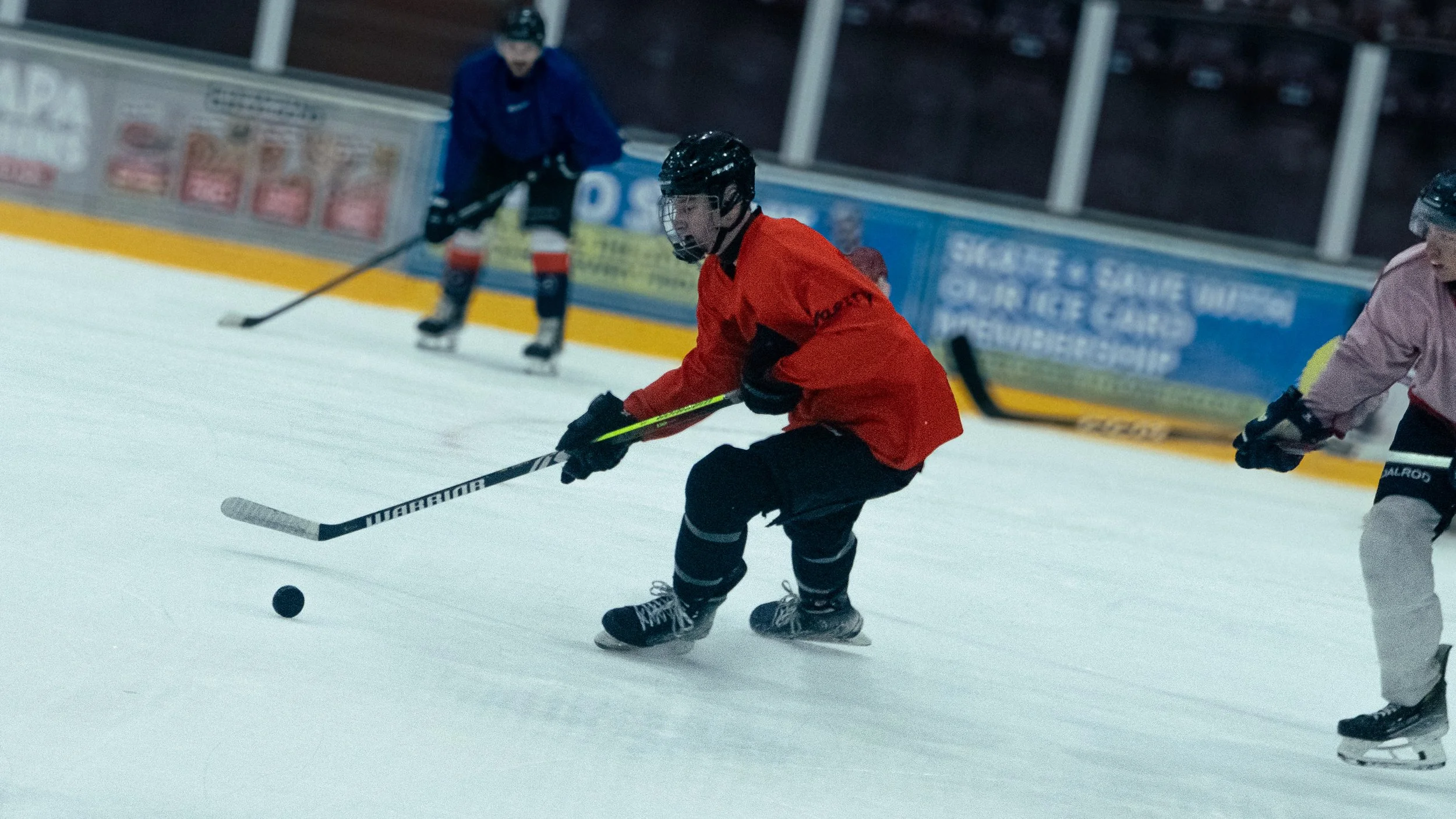 Young hockey players on ice rink, one in red jersey controlling puck with hockey stick, other in pink jersey skating nearby, and a third player in blue in the background.