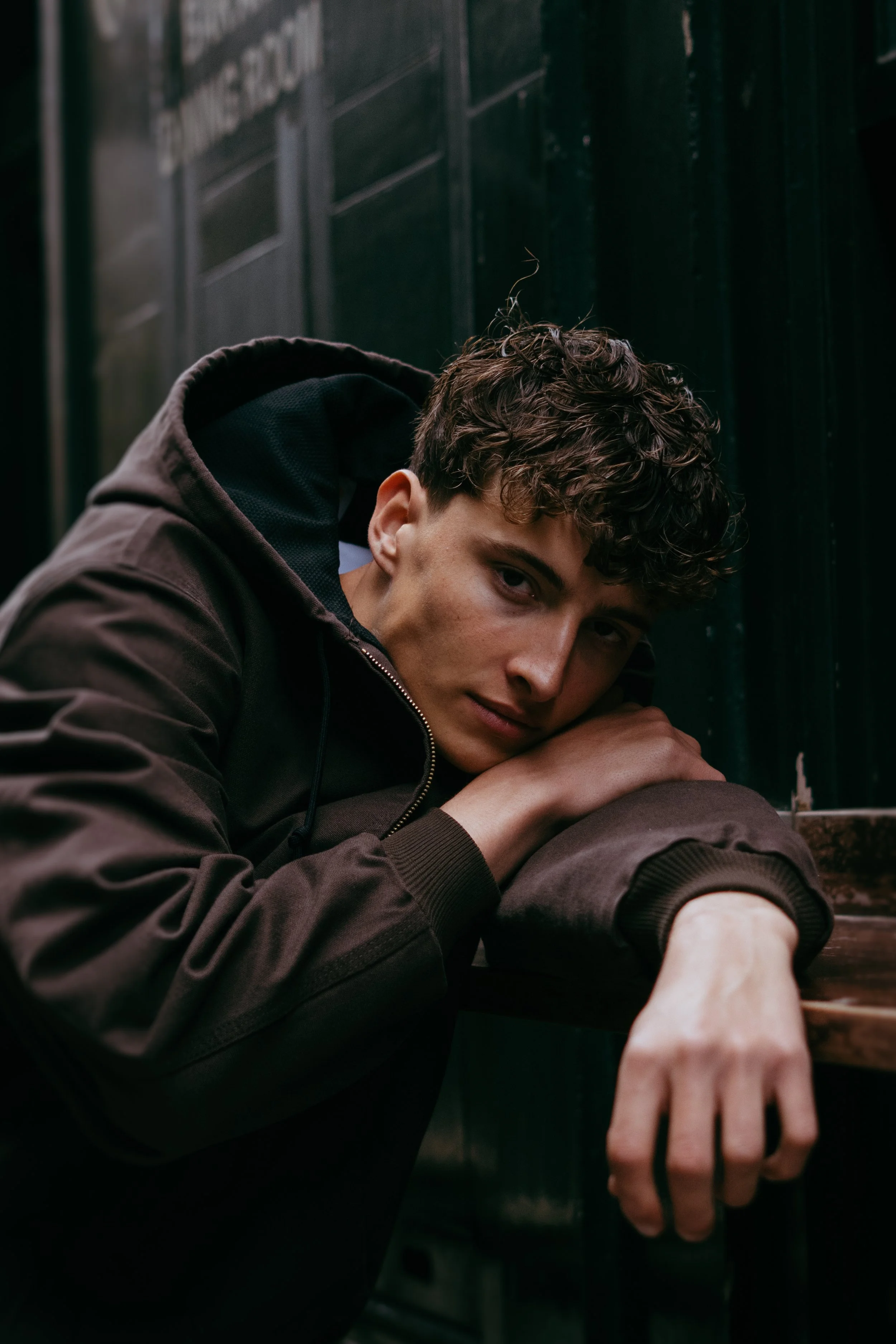 Young man lying down with head resting on arms, wearing a dark jacket, in a moody, dimly lit setting.