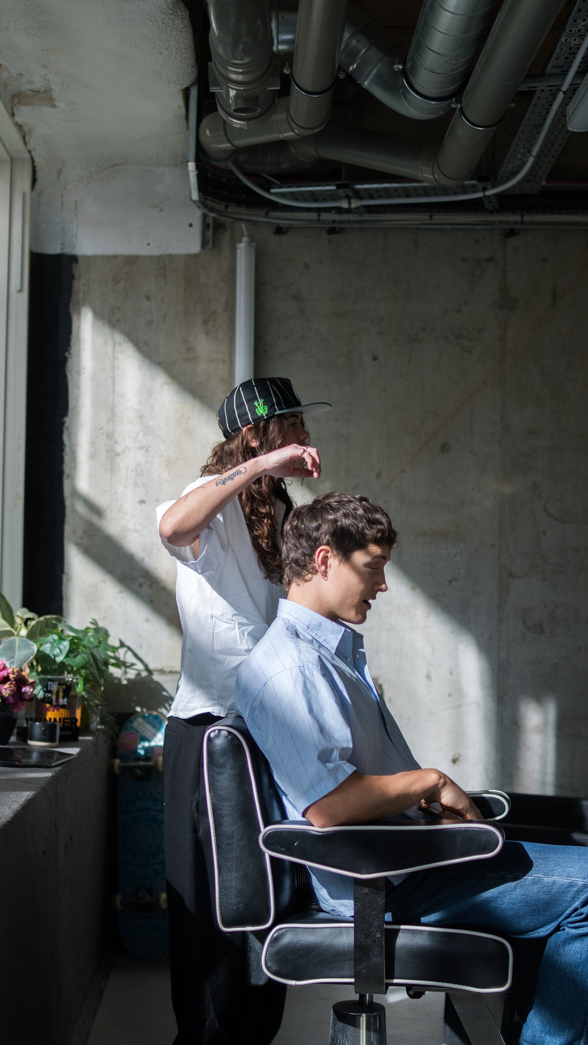 A woman getting a haircut in a modern barber shop, with exposed pipes on the ceiling and plants on a nearby counter.