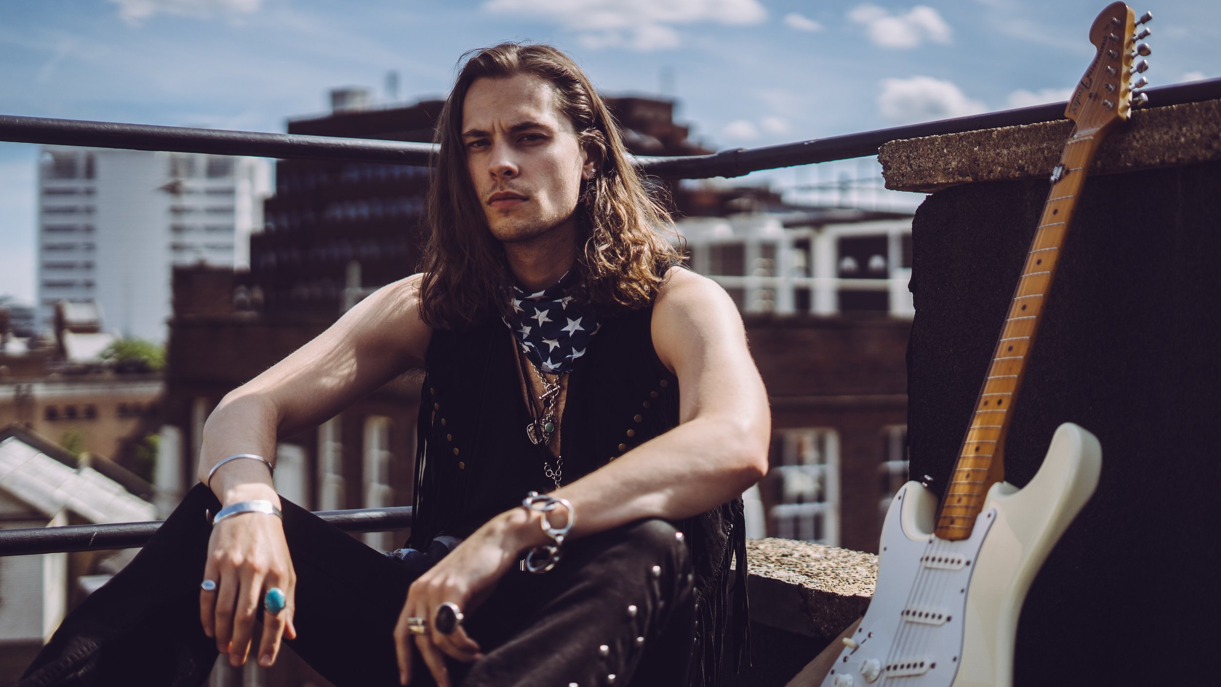 A young man with long hair, wearing a sleeveless black vest, sitting on a rooftop with a white electric guitar beside him, looking at the camera with a serious expression against a background of city buildings and blue sky.