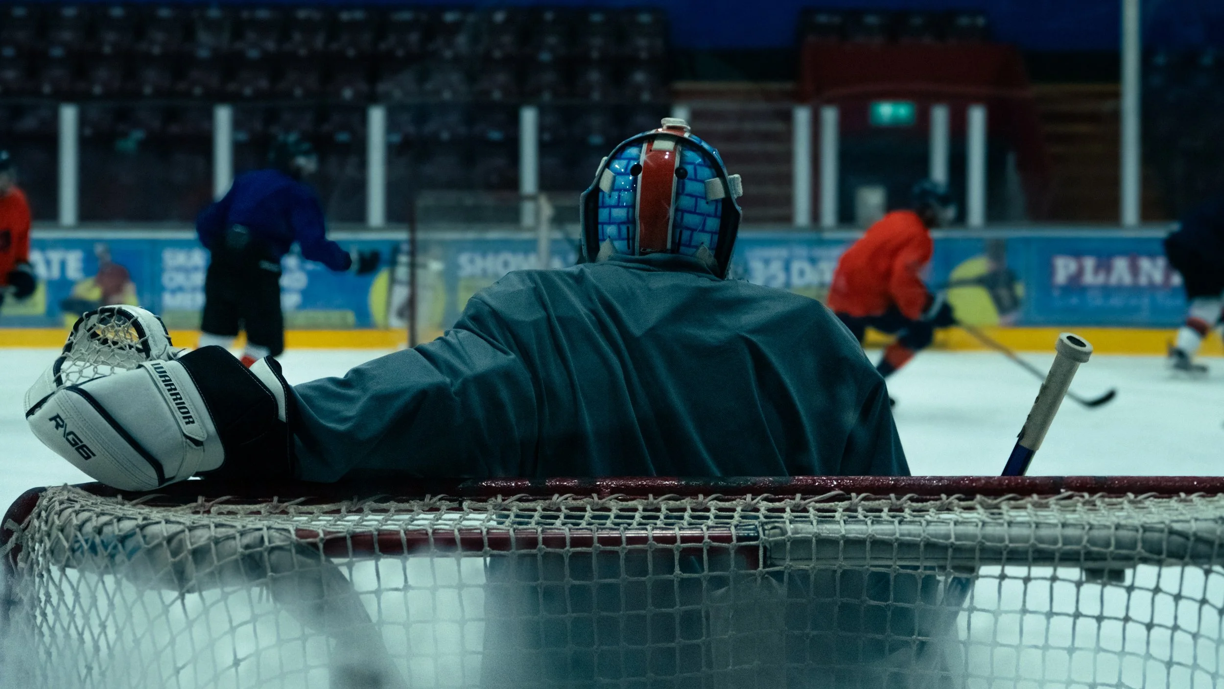 Hockey goalie in black jersey and helmet watching players on ice during practice at an indoor rink.