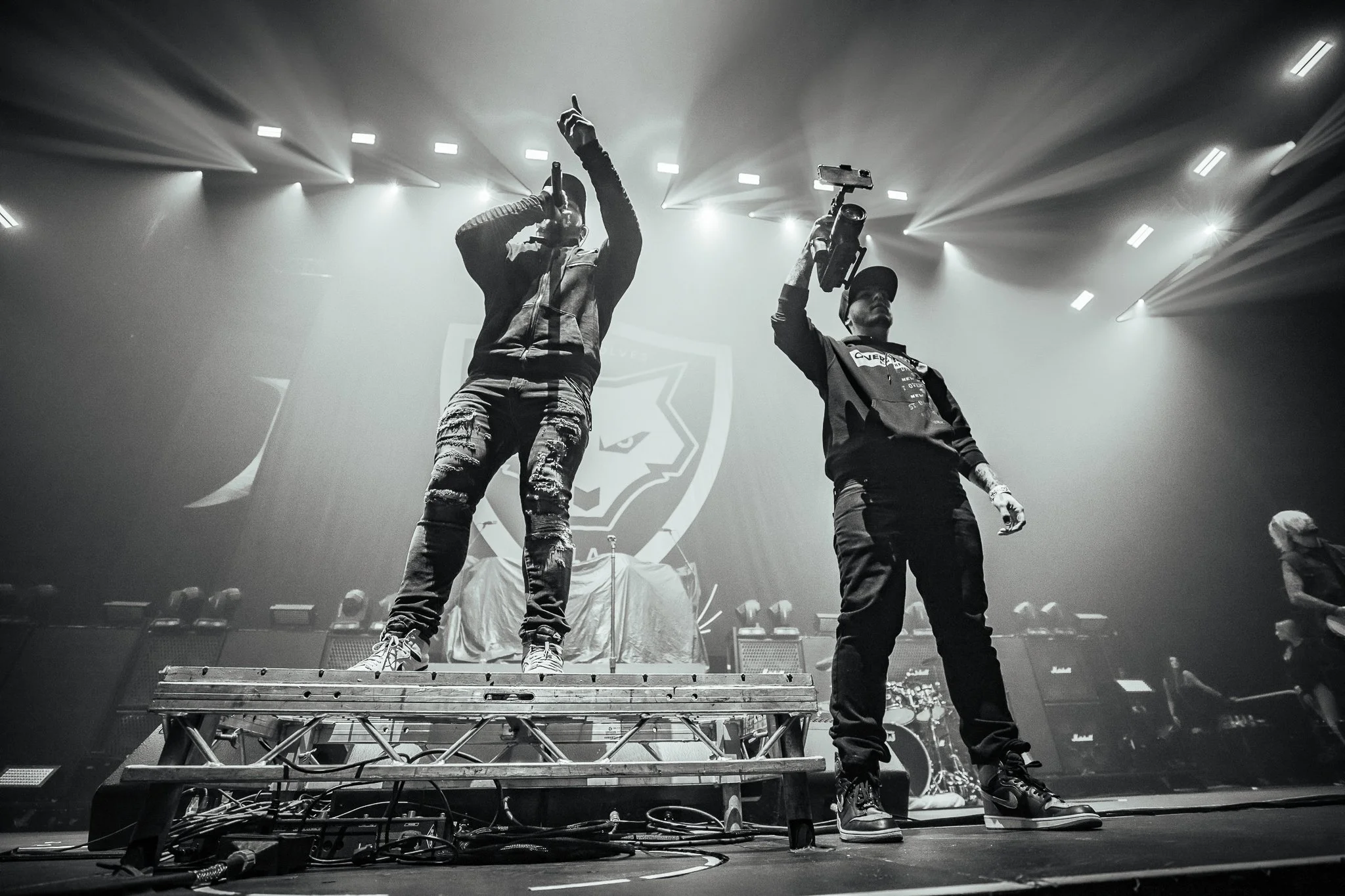 Two performers on stage, one holding a microphone and pointing upward, the other holding a microphone and a camera, with stage lights and a logo of a wolf's head projected in the background.