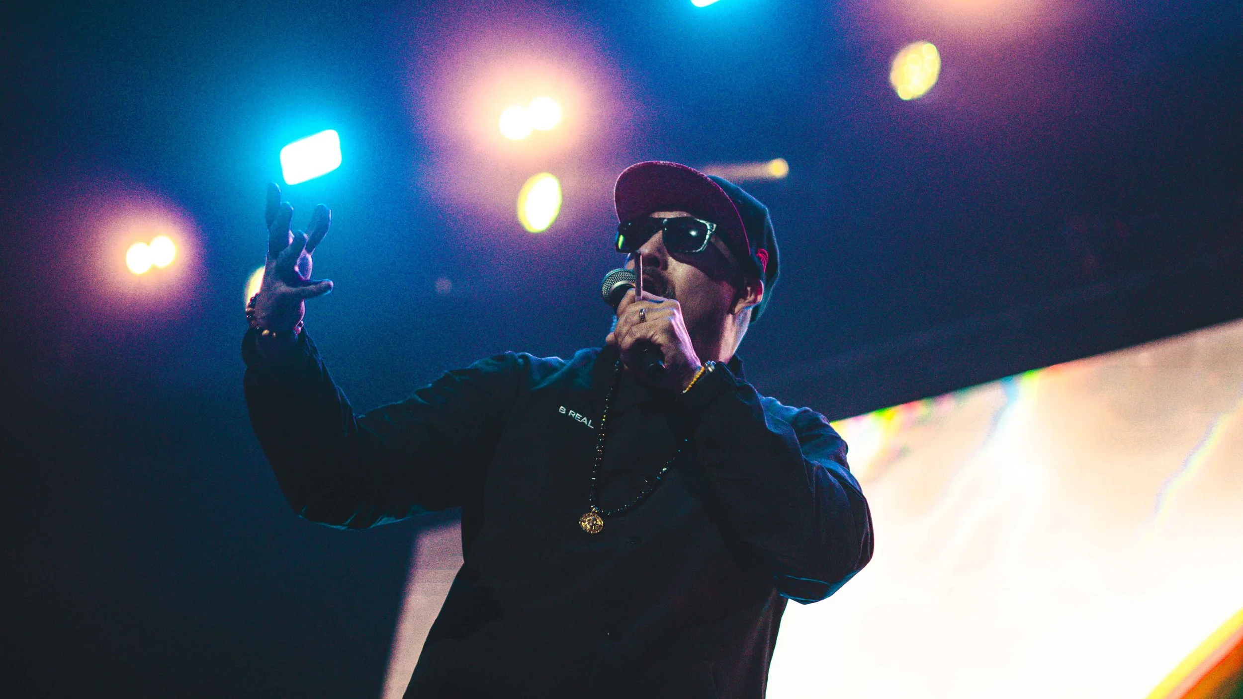 A male performer on stage, wearing sunglasses, a black cap, and a black jacket, holding a microphone and singing under colorful stage lights.