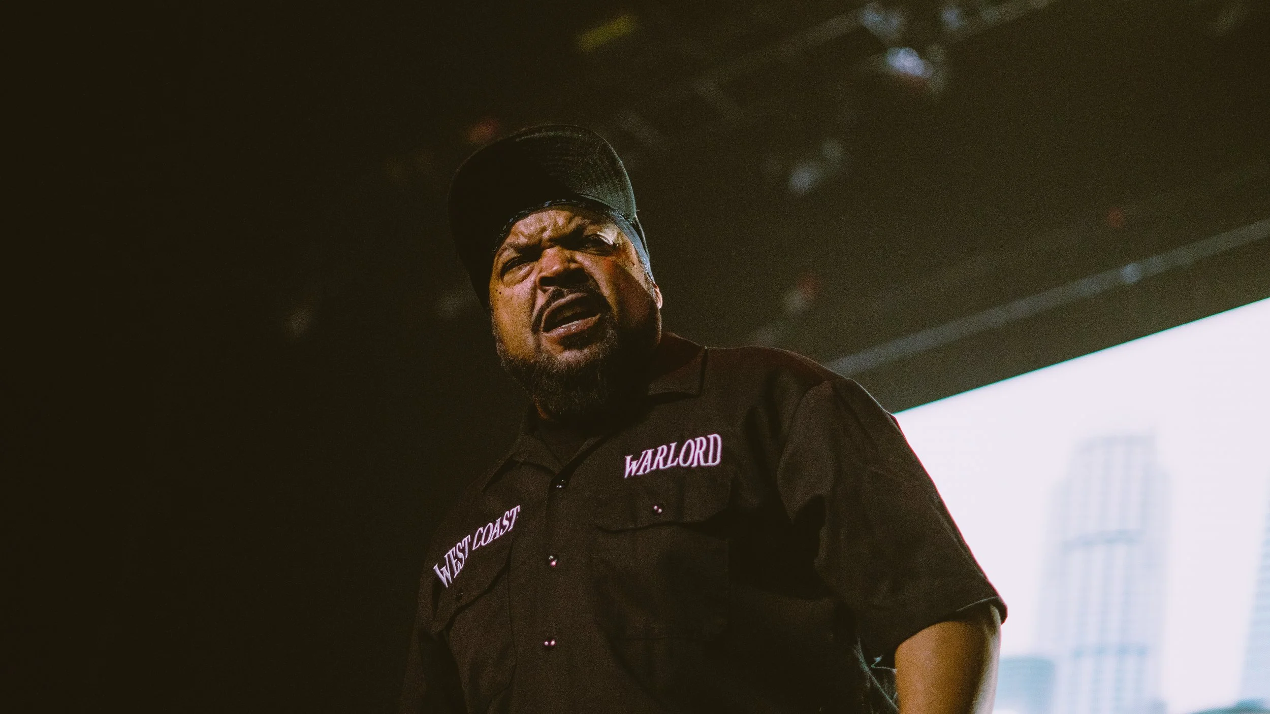 Man with a black cap and dark shirt with patches reading 'Warlord' and 'West Coast' looking confused or angry, standing indoors with city buildings visible through a window behind him.