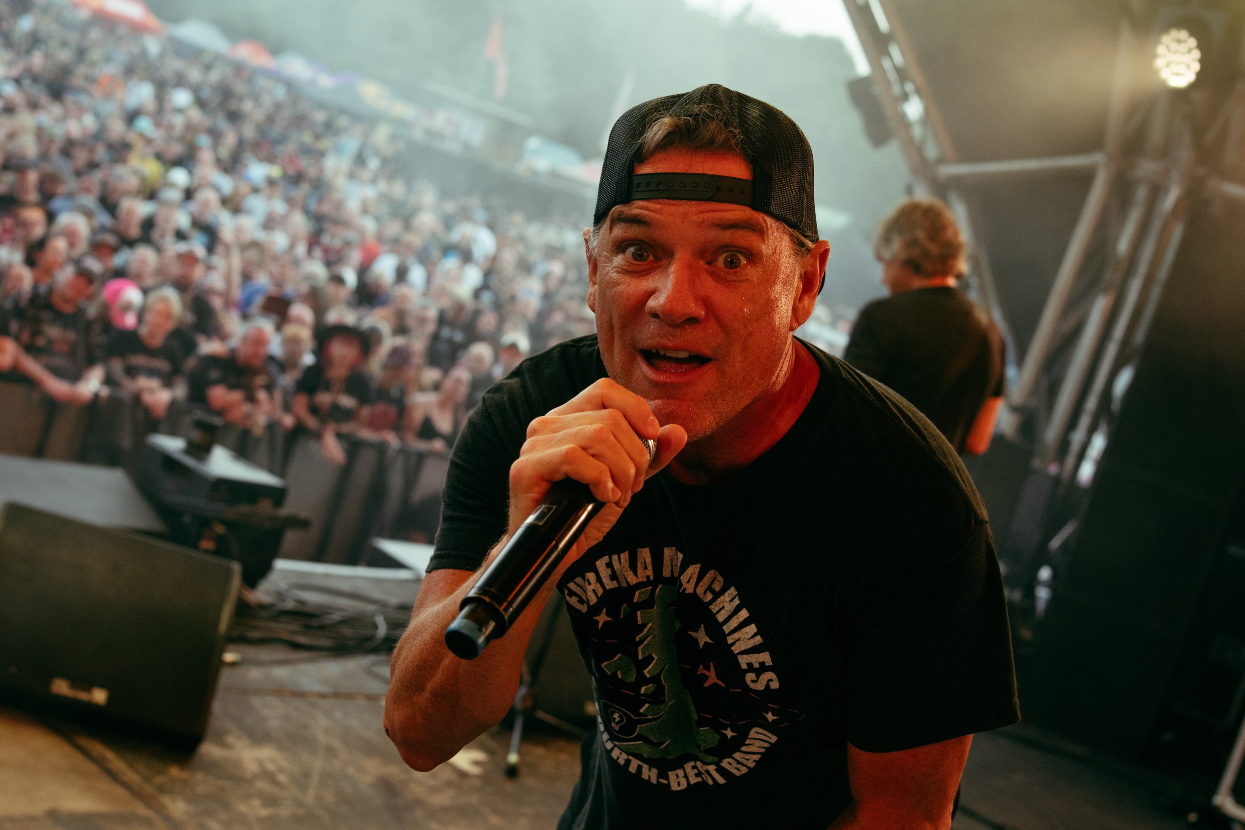 A performer in a black t-shirt and baseball cap holding a microphone on stage at a concert, with a large crowd of people in the background.