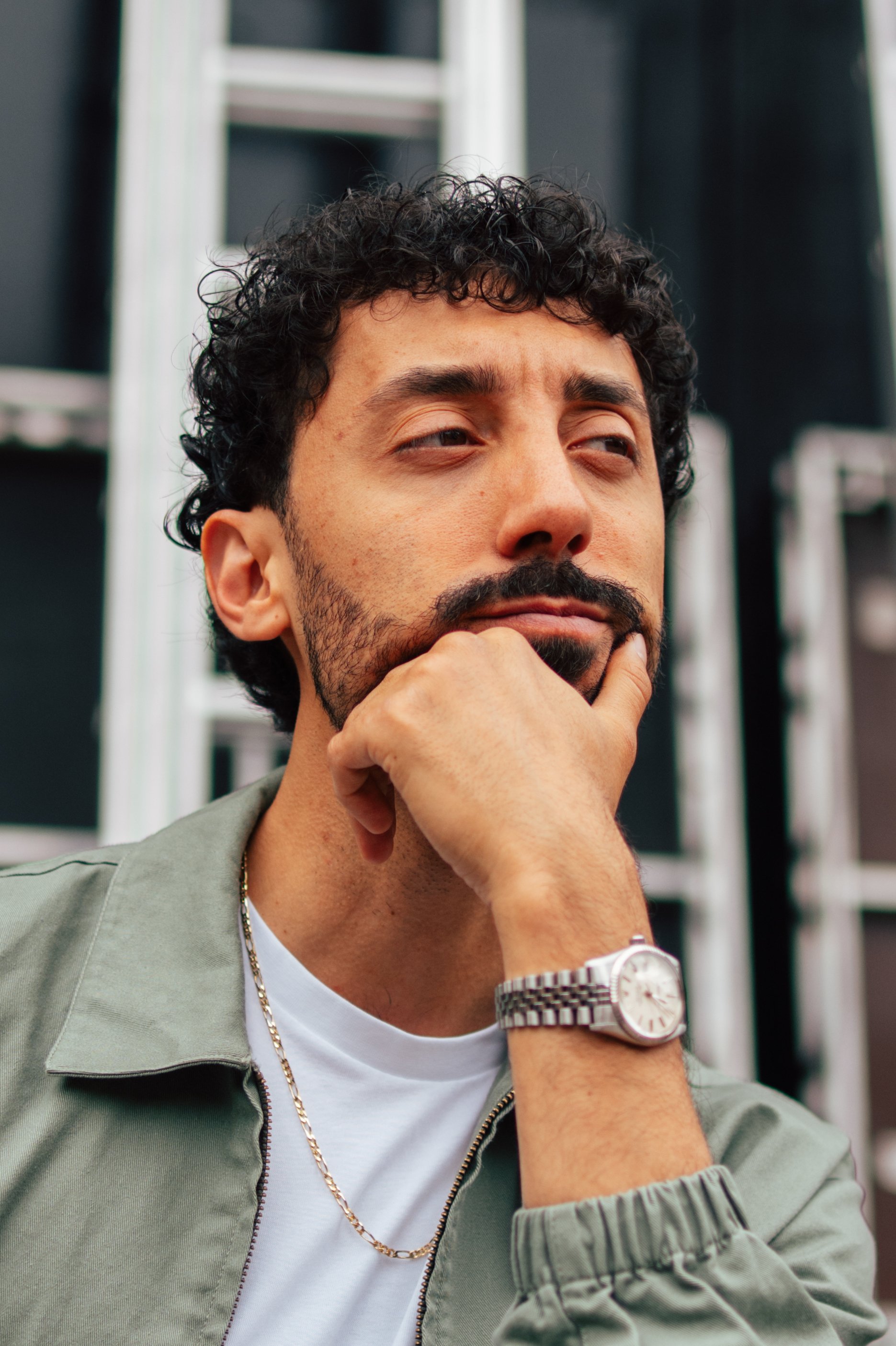 A man with curly dark hair and a beard is resting his chin on his hand, looking thoughtful. He is wearing a green jacket, a white t-shirt, a silver watch, and a gold chain necklace. The background consists of black panels and a white ladder.