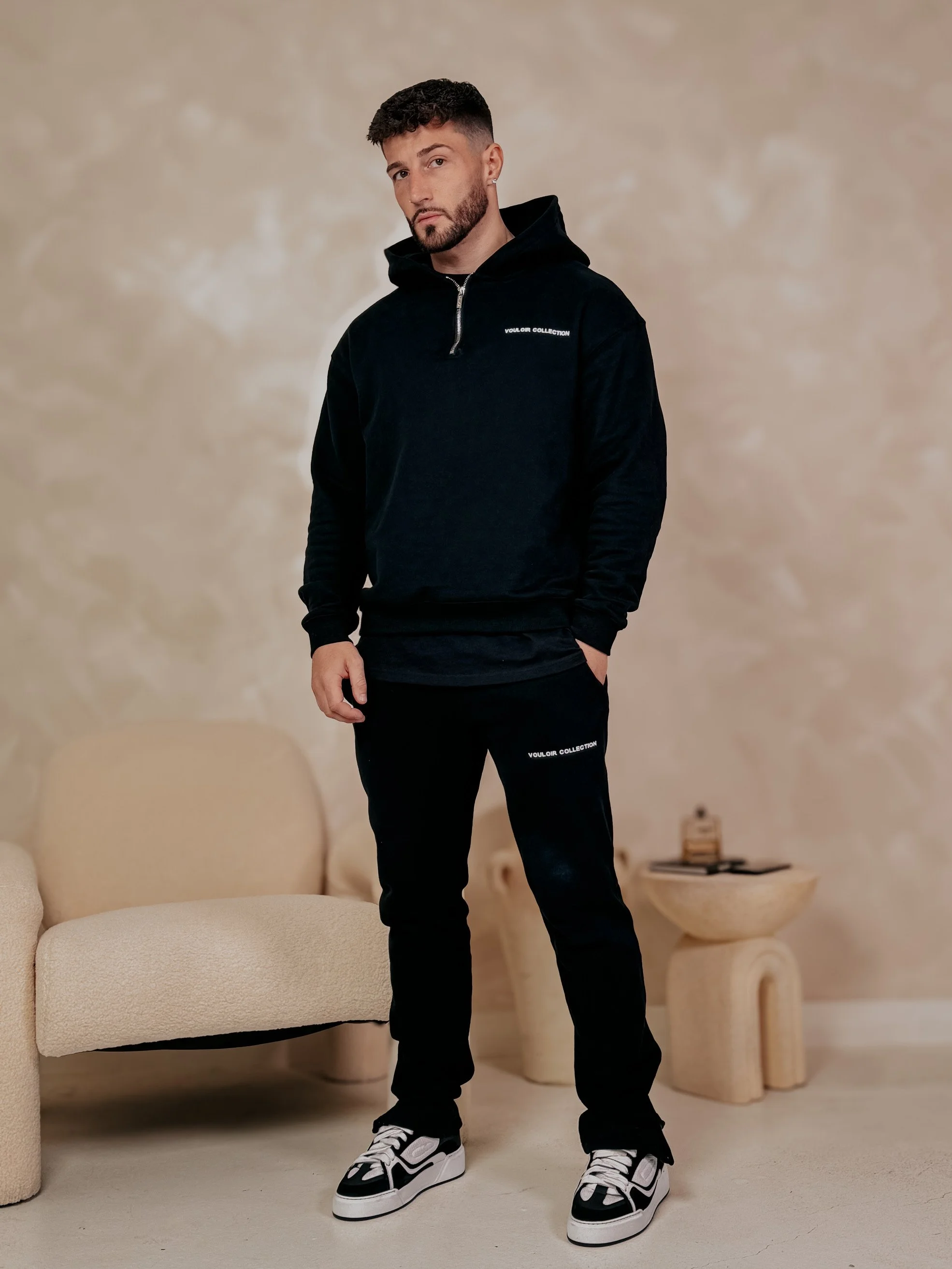 Young man with dark hair and a beard standing in a room with beige textured walls, wearing a black hoodie and matching black sweatpants with white lettering, and black and white sneakers.