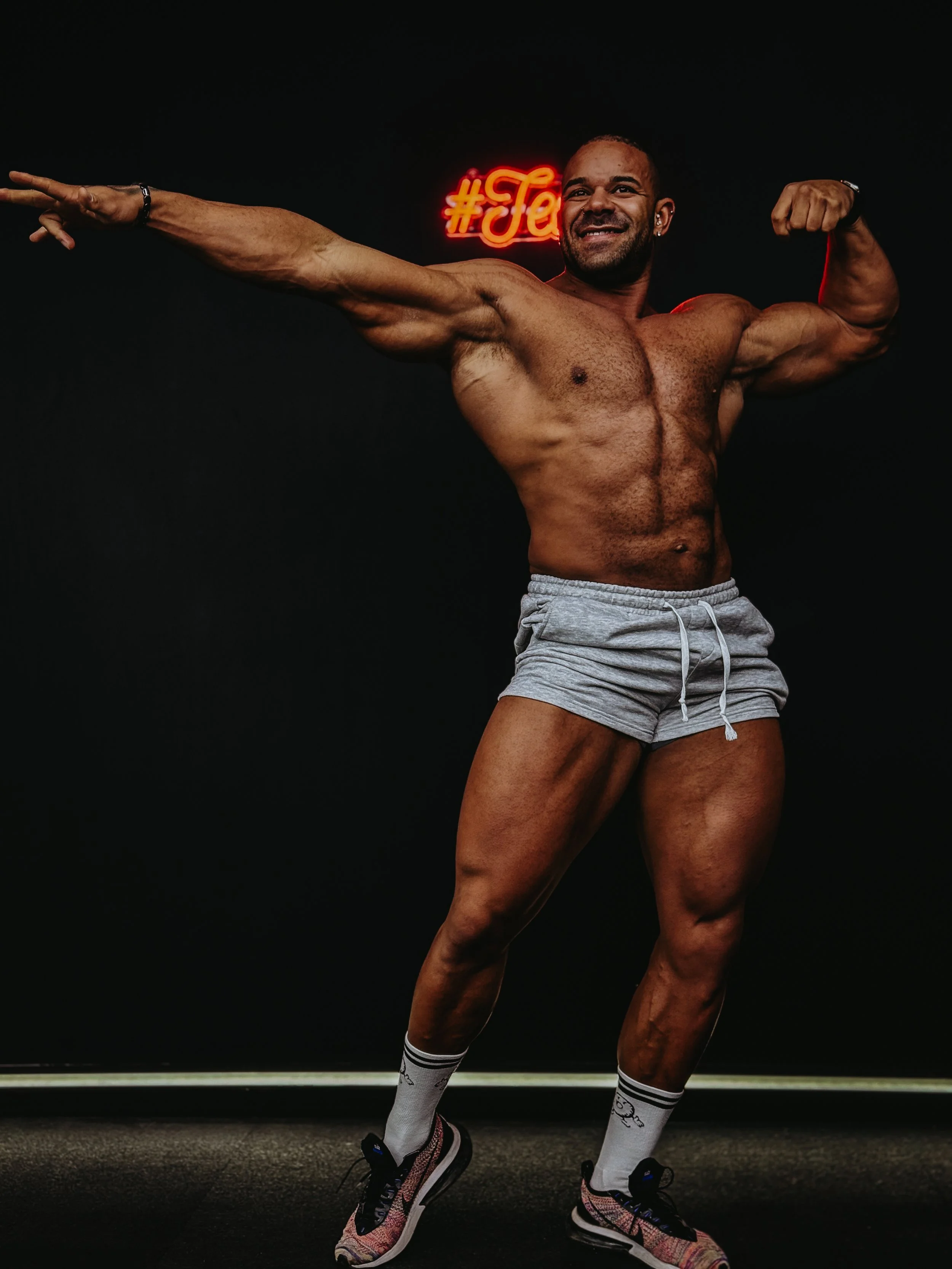A muscular man with a beard posing with his right arm extended and left arm flexed, shirtless, wearing gray shorts, running shoes, and socks, standing against a black background with a red neon sign reading '#Jee' behind him.