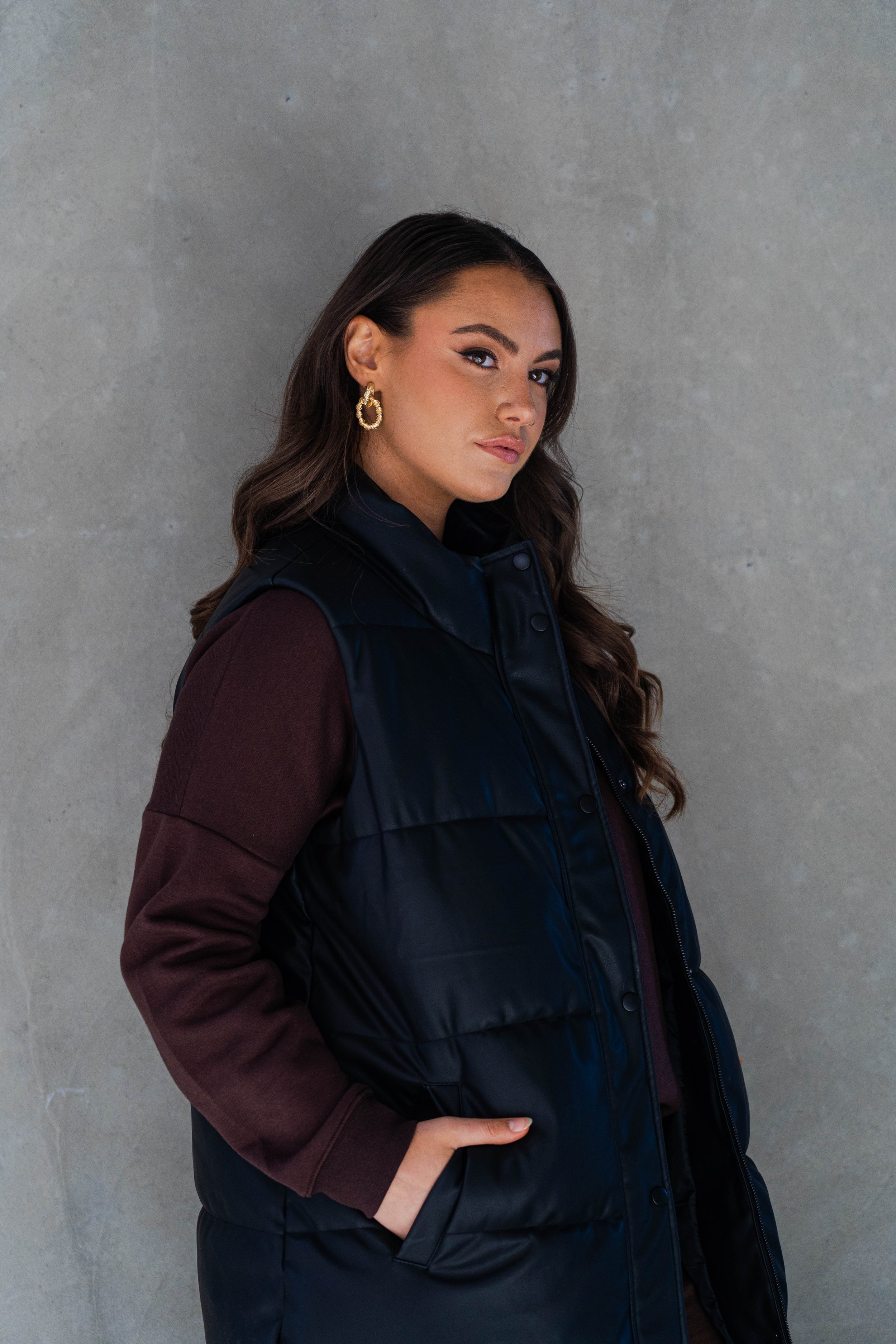A woman with dark brown hair and gold hoop earrings stands against a plain grey wall, wearing a dark puffy vest over a maroon long-sleeve shirt, with her hand in her pocket.