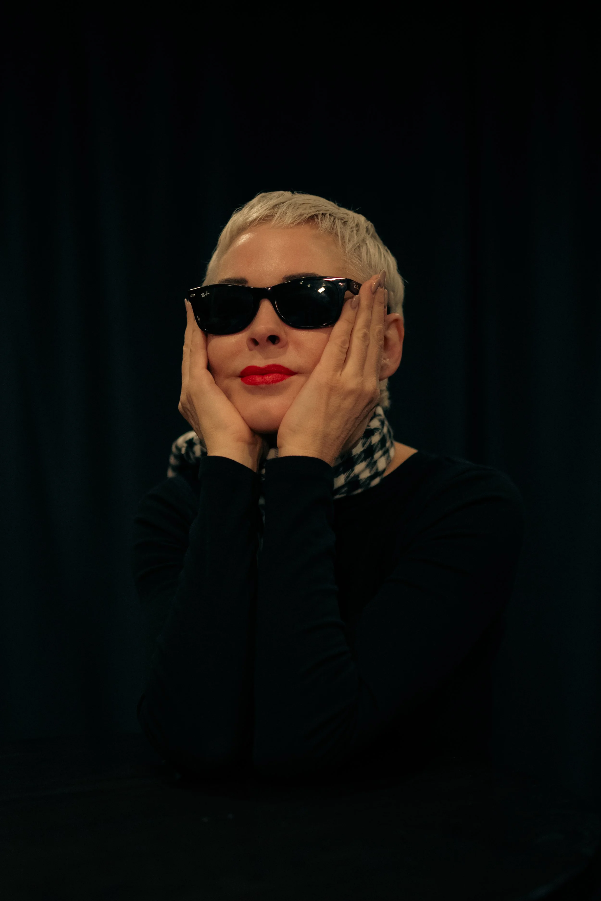 Woman with platinum blonde hair, wearing sunglasses, red lipstick, and a black outfit, resting her face in her hands against a dark background.