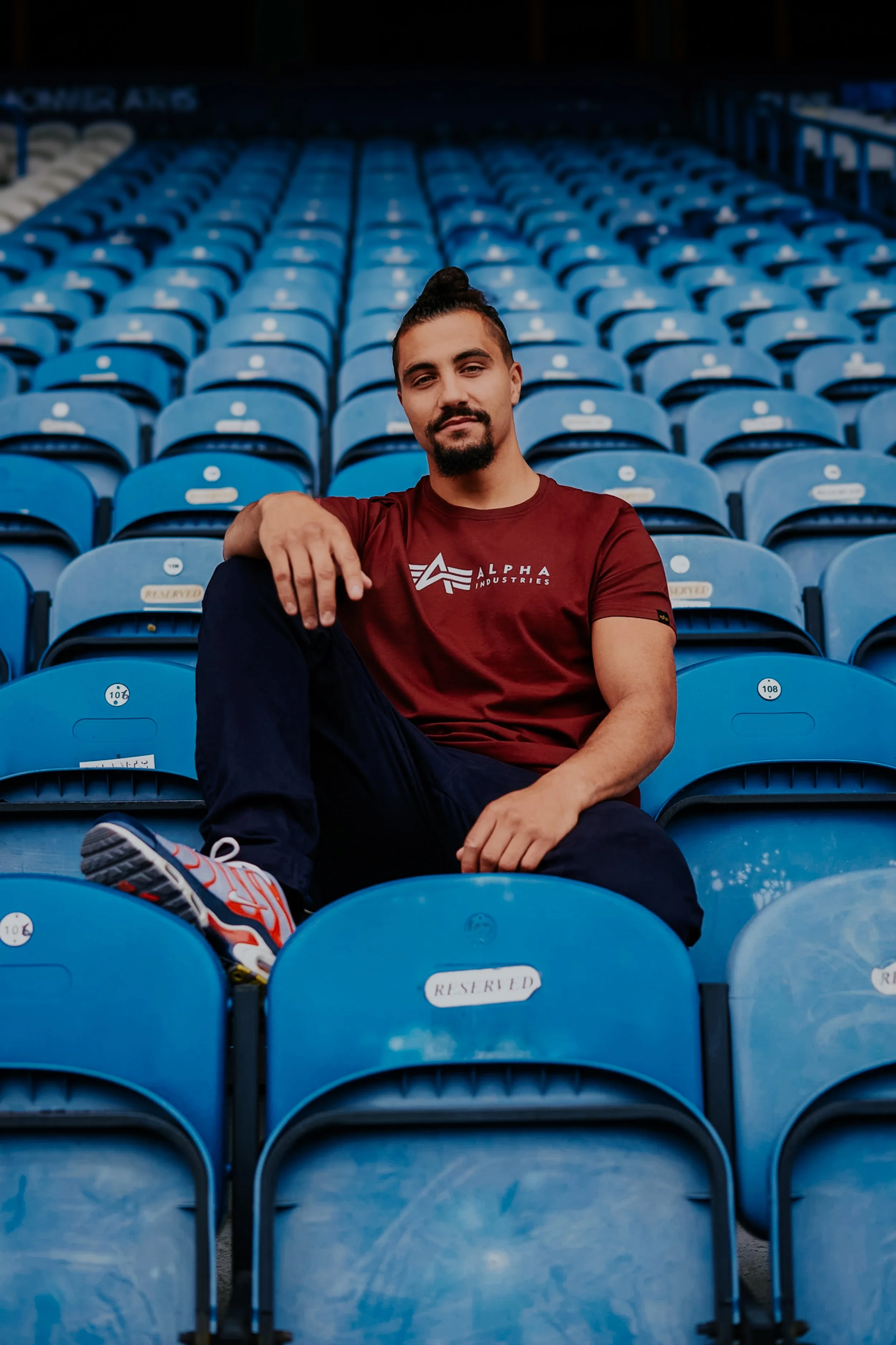 A man sitting on blue stadium seats, wearing a maroon T-shirt and sneakers, with a confident expression in an empty stadium.