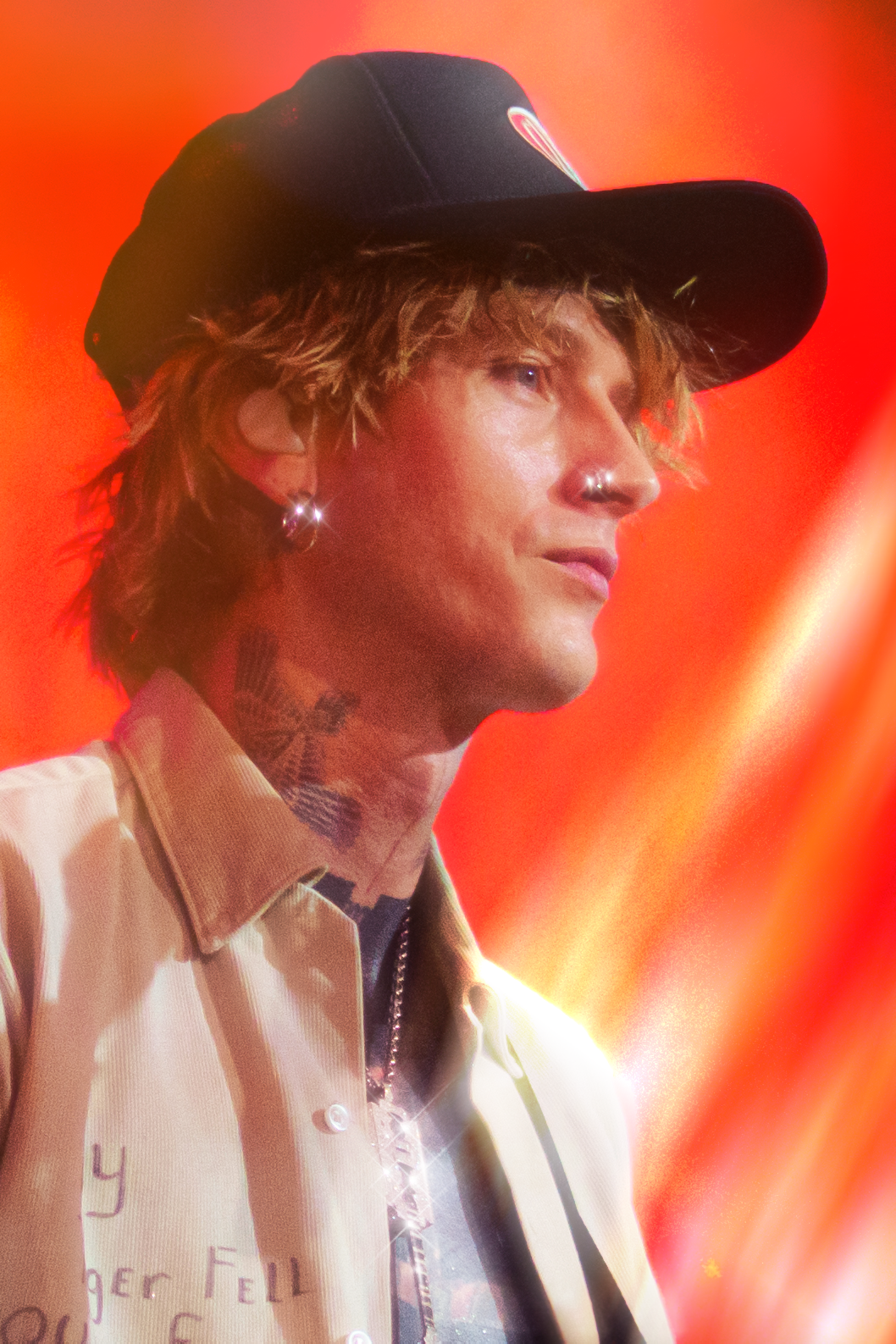 A young man with blond curly hair wearing a black baseball cap and earrings. He has tattoos on his neck and is dressed in a beige shirt with writing on it. The background is bright orange with light effects.