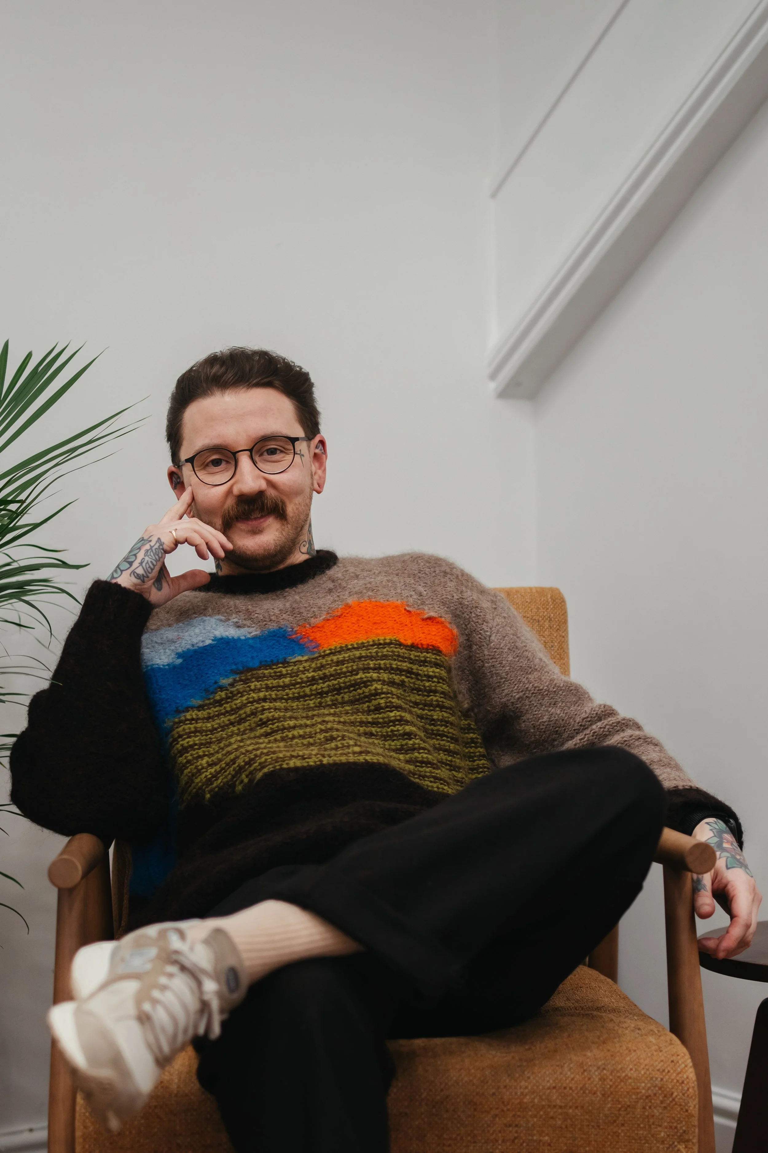 A man with glasses, a mustache, and tattoos wearing a colorful sweater, sitting in a chair indoors, smiling at the camera with one hand resting on his face.