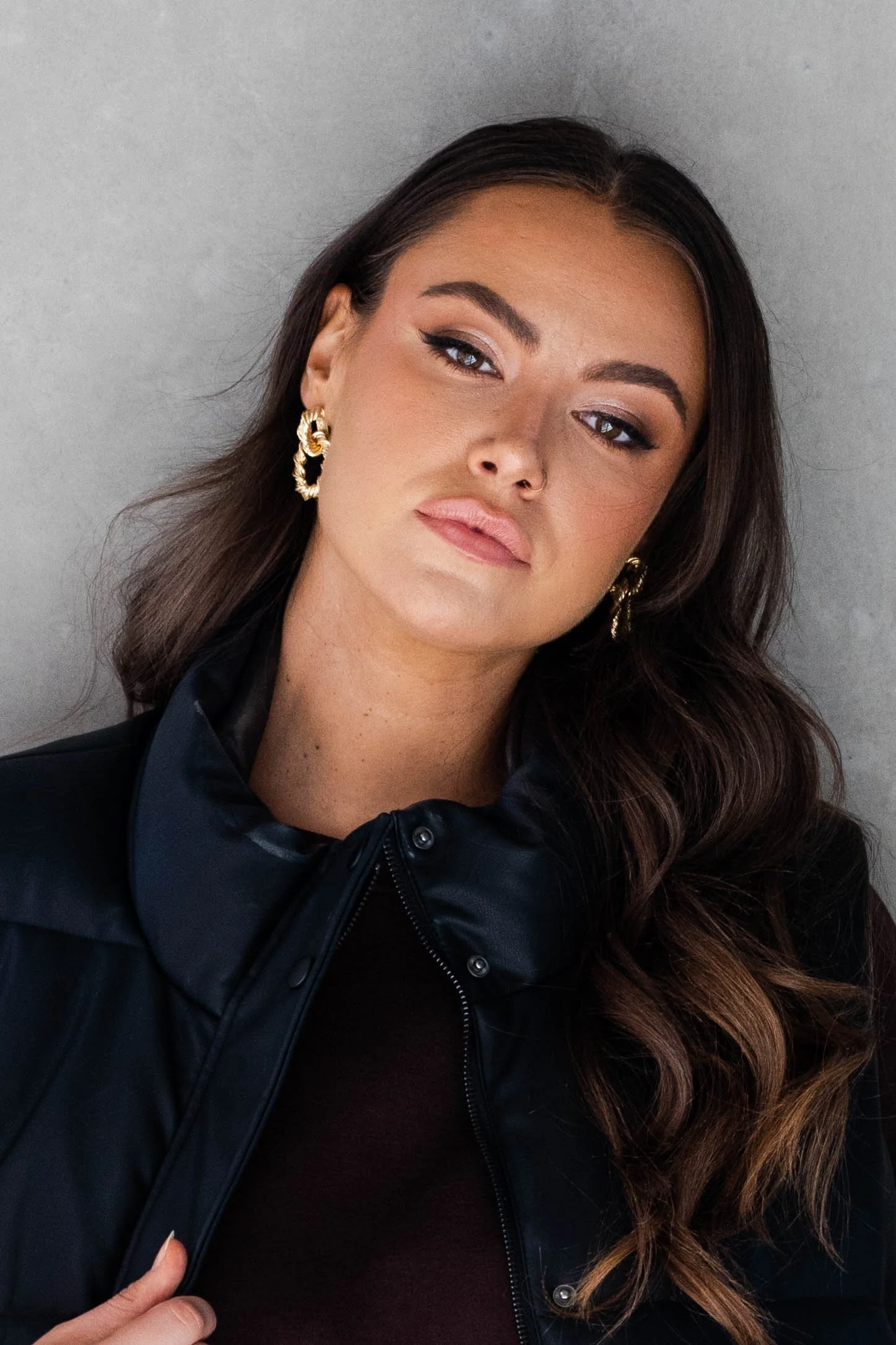 A young woman with long wavy brown hair, wearing gold earrings and a black leather jacket, posed against a plain gray background.