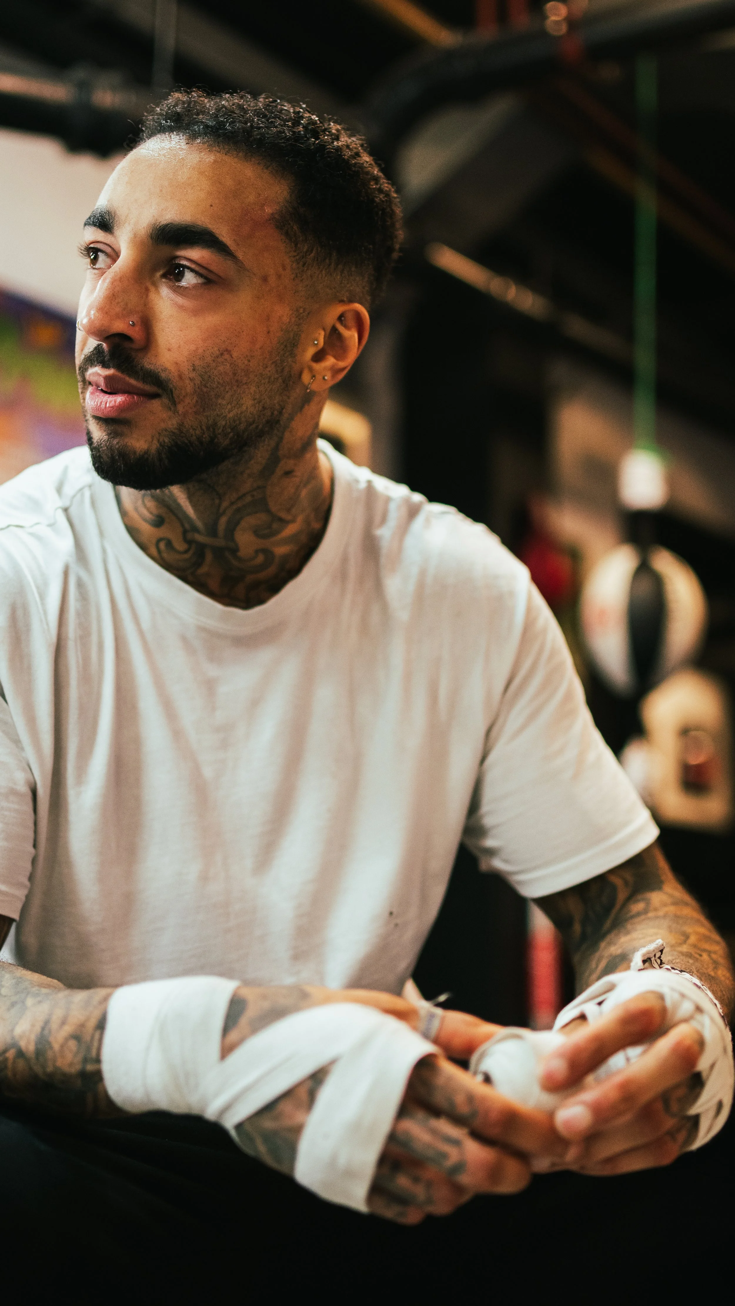 A tattooed man in a white T-shirt sitting in a gym or locker room, holding his wrapped hands.