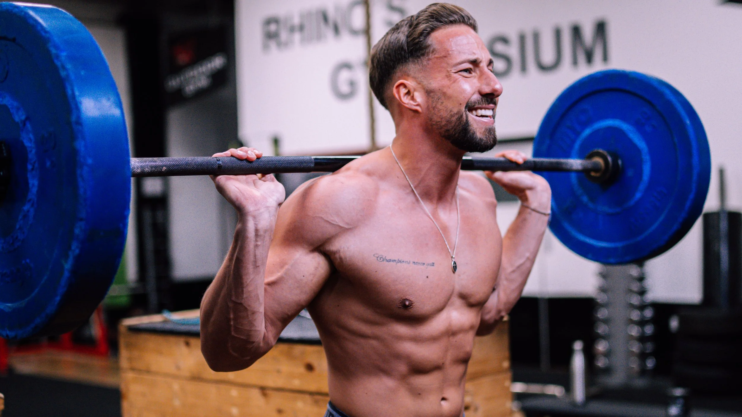 A shirtless man with tattoos lifting a weighted barbell on his shoulders at the gym, smiling and showing effort.