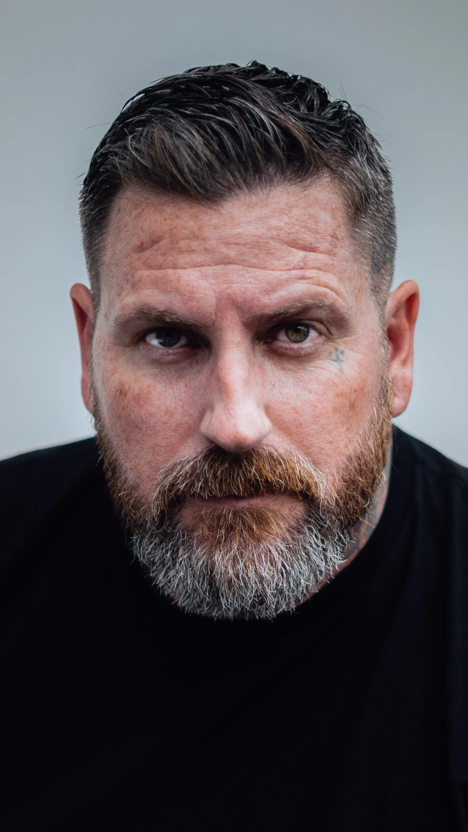 Close-up portrait of a middle-aged man with dark hair, beard, and mustache, looking directly into the camera with gray eyes, wearing a black shirt against a plain background.