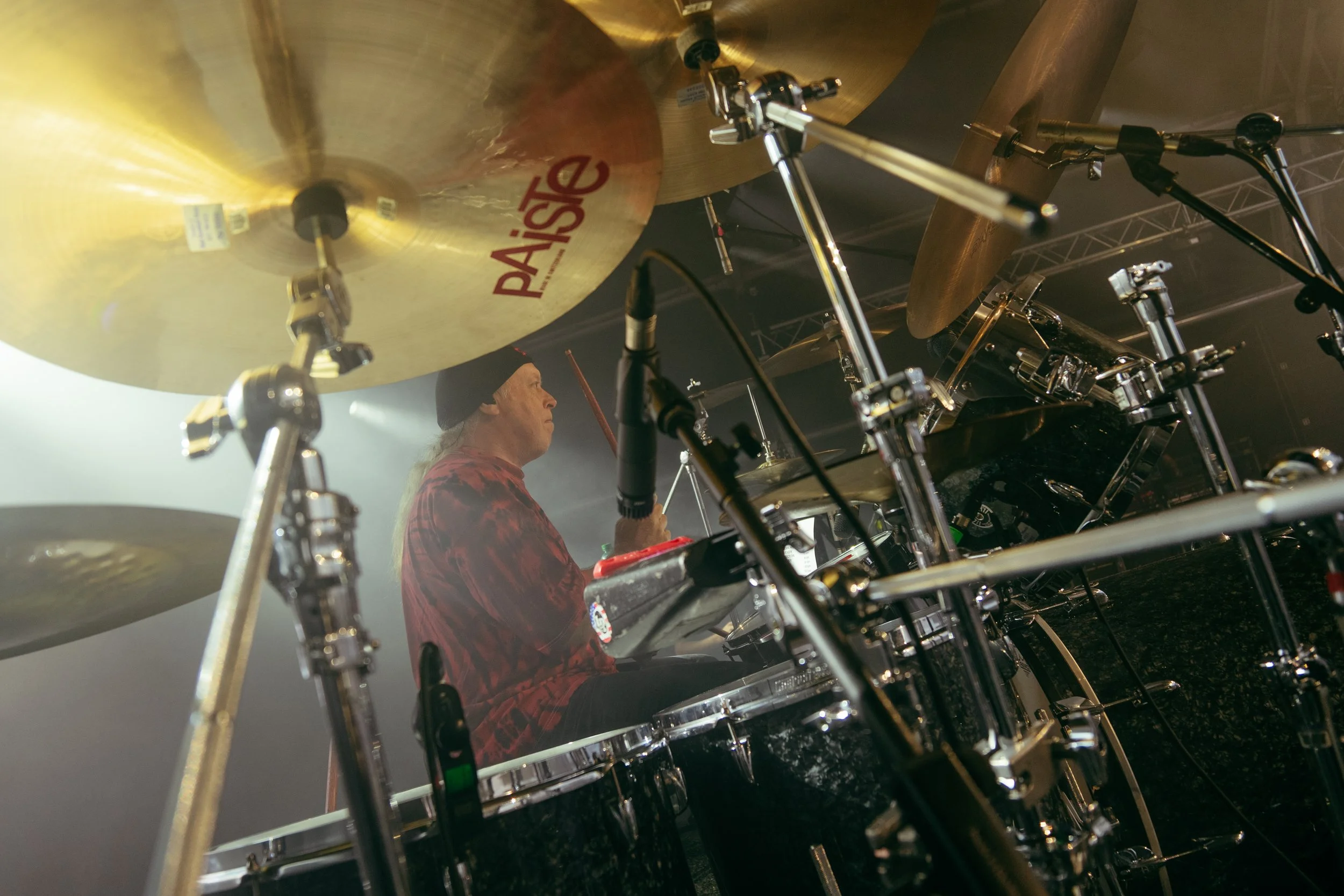 A drummer in a black beanie and red shirt playing a drum set on stage, viewed from a low angle with cymbals in the foreground.