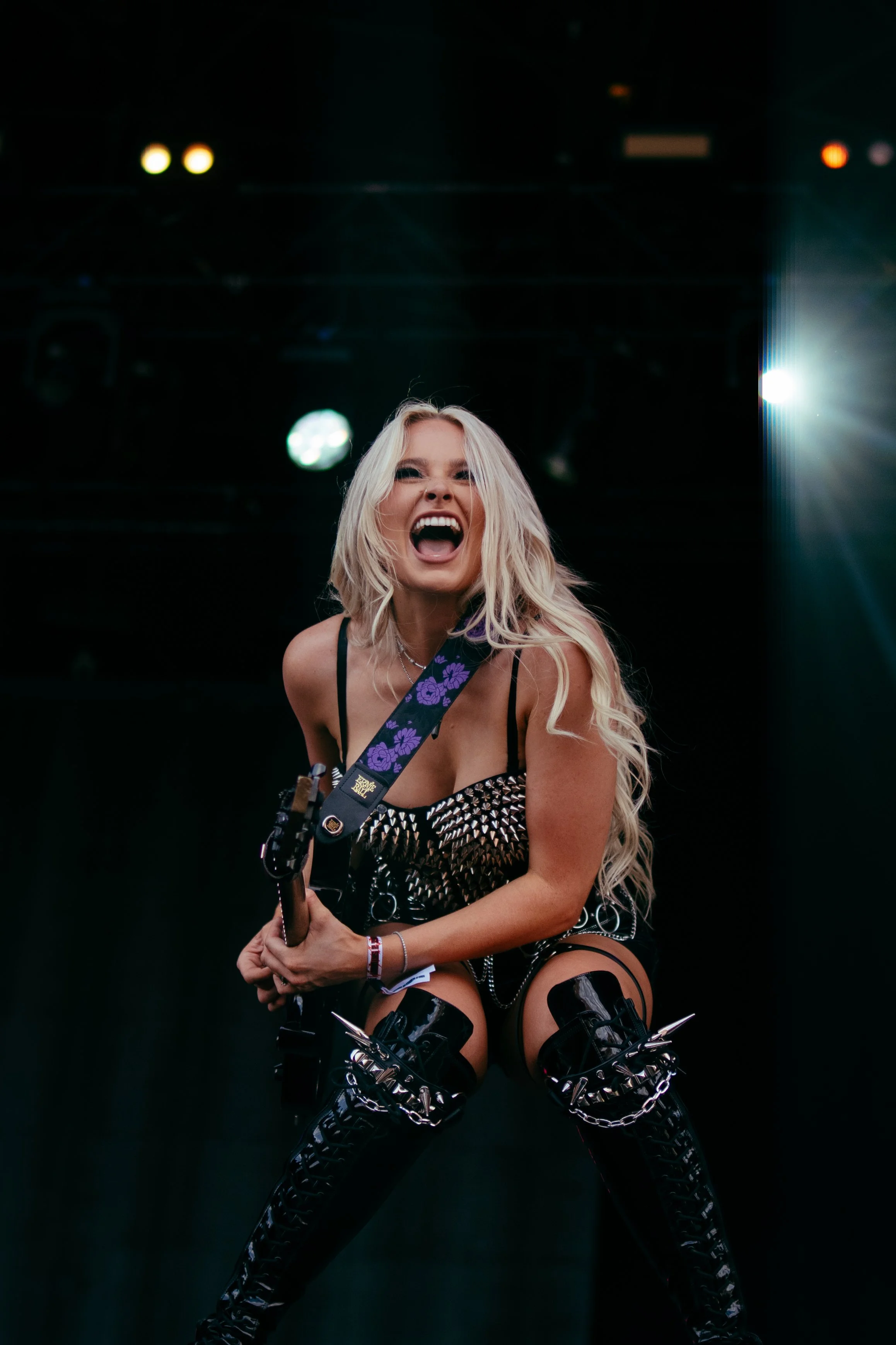 A woman with long blonde hair and a black studded outfit performing on stage, holding an electric guitar with a purple strap, with a dark background and stage lights.